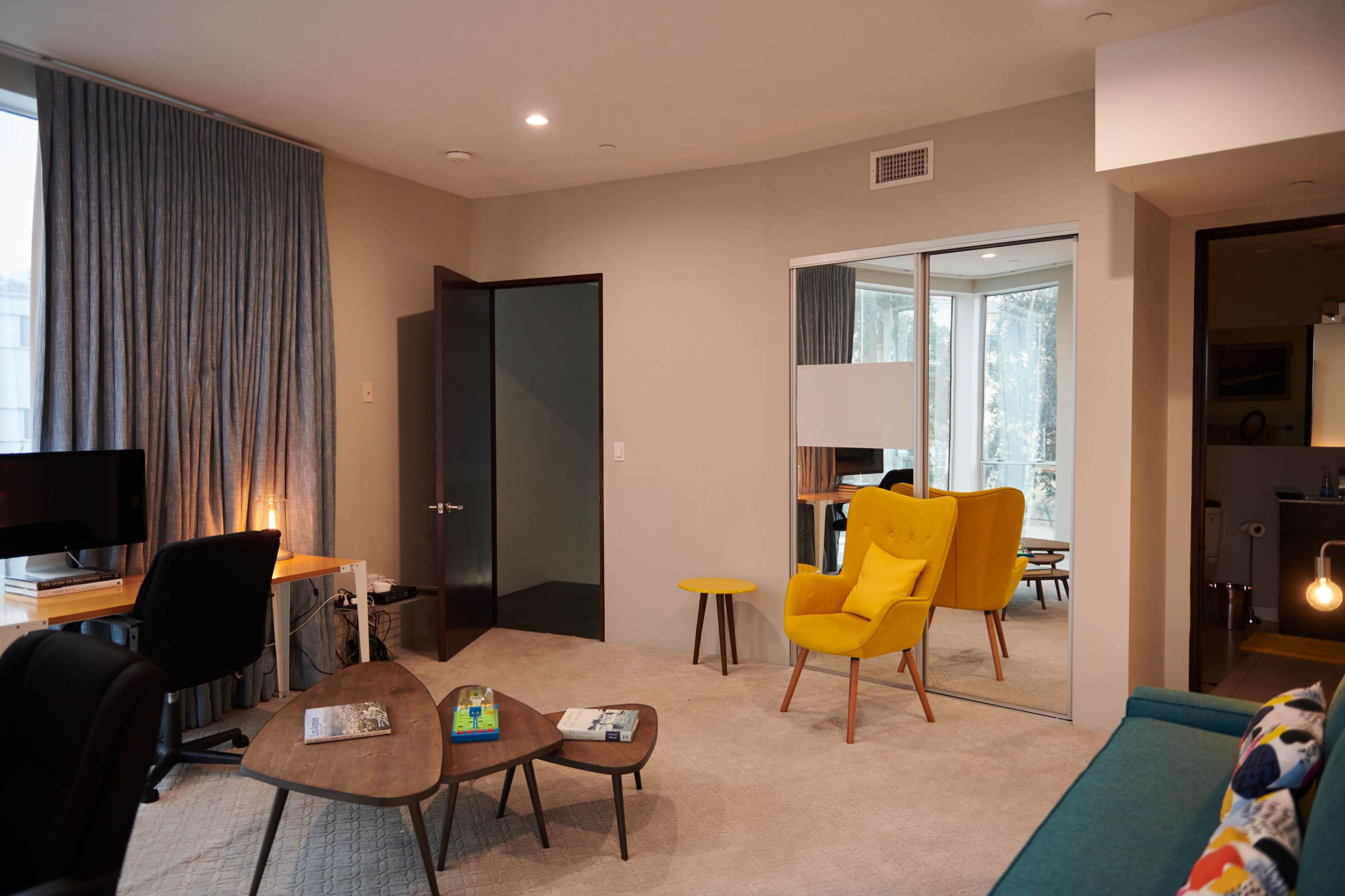 A modern living room featuring a yellow armchair, a small coffee table, and a work desk beside a window with blue curtains.