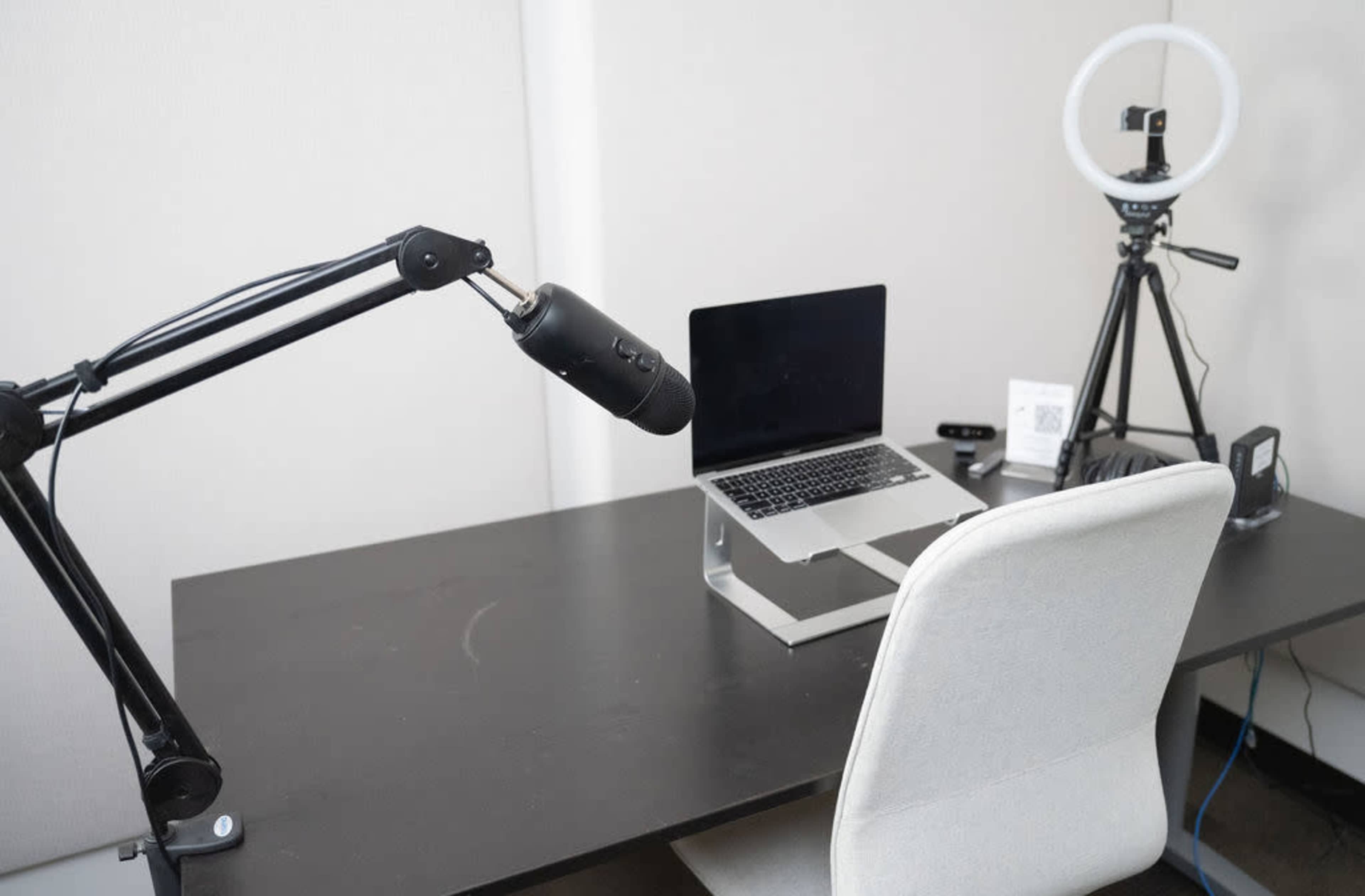 A desk setup features a laptop on a stand, a microphone mounted on an adjustable arm, a ring light, and a tripod, with a modern chair positioned in front.