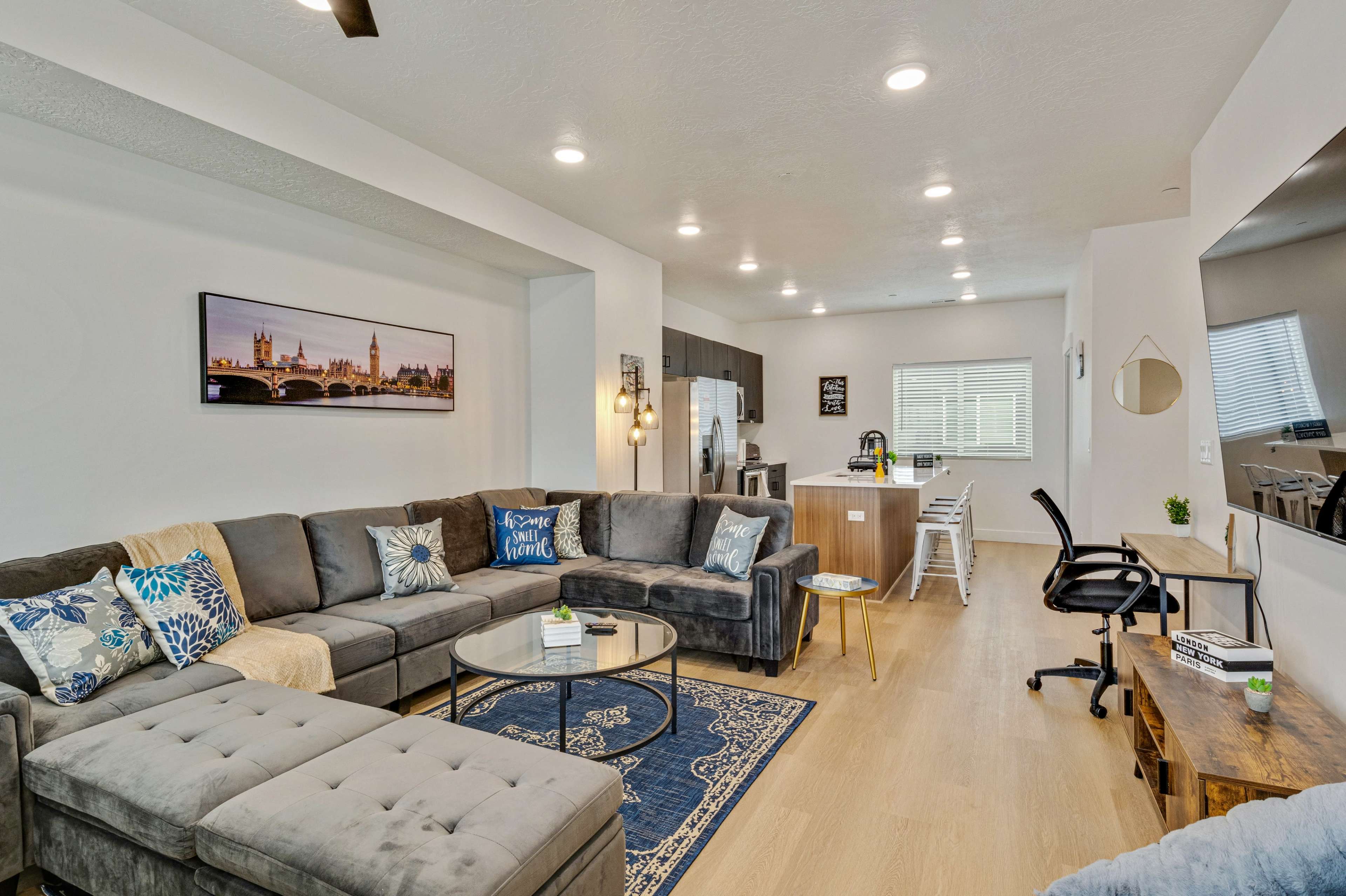 A modern living room with a gray sectional sofa, a round glass coffee table, and a kitchen area in the background.