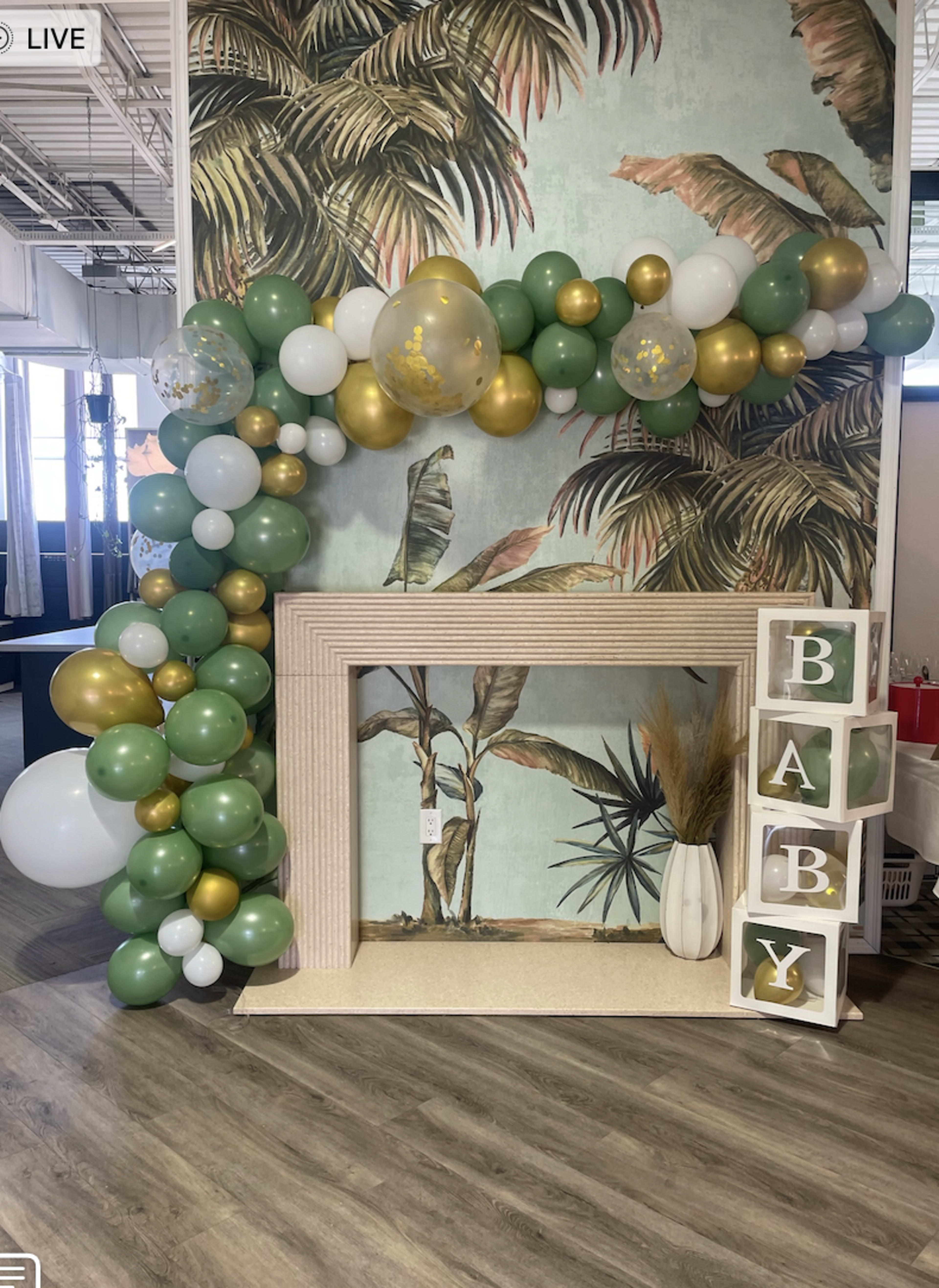 A decorative display featuring a balloon arch in green, gold, and white, alongside large blocks spelling "BABY," set against a tropical-themed wall and a fireplace.