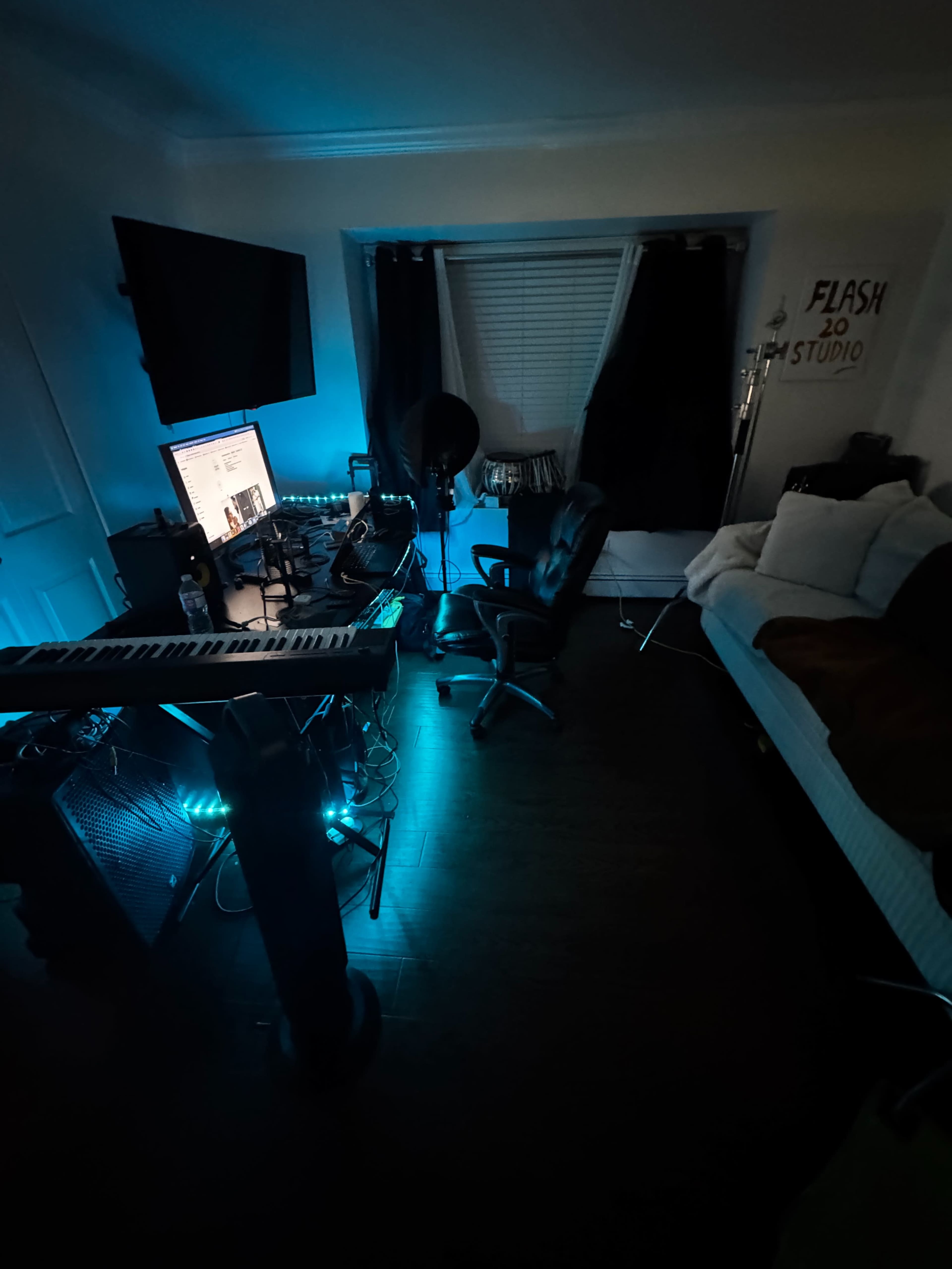 The image shows a dimly lit room featuring a computer setup, a keyboard, and a bed, with blue lighting casting a cool tone throughout the space.