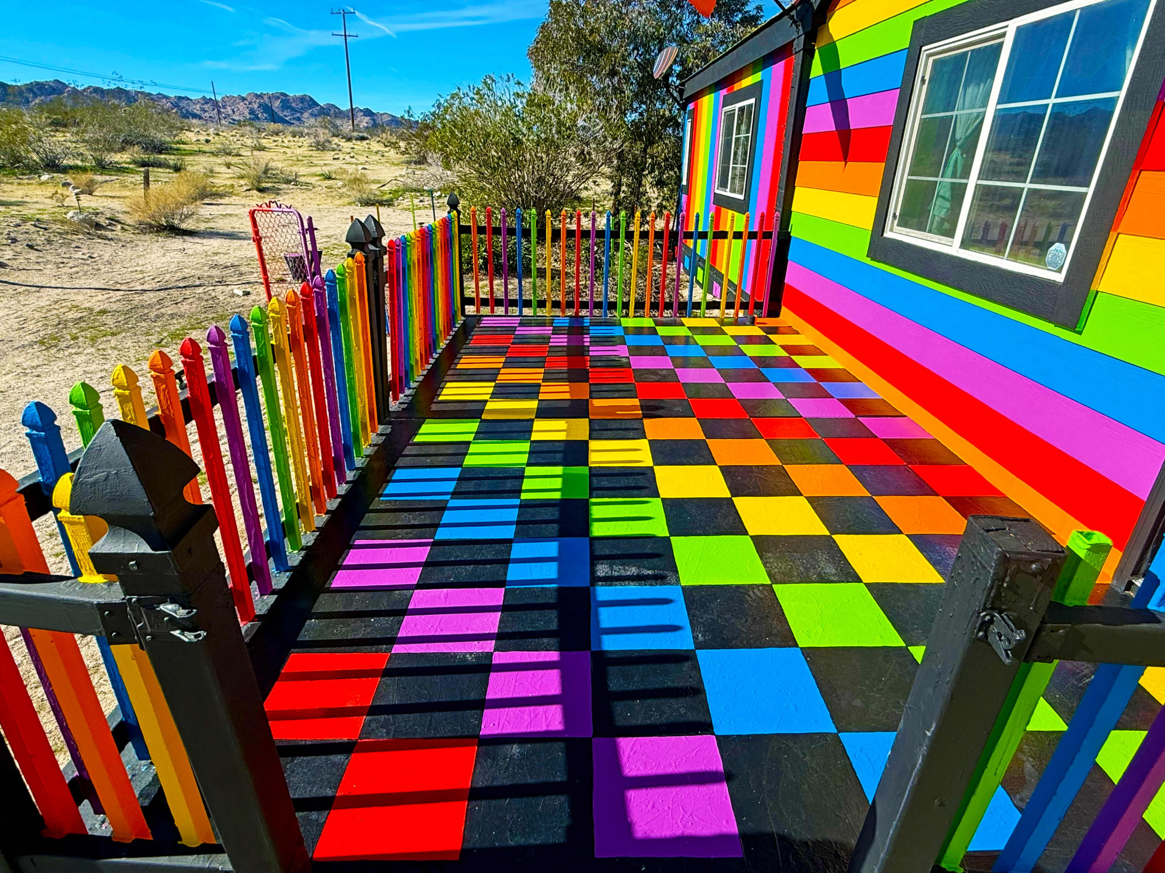 The image shows a brightly painted outdoor area with a checkered floor in vibrant colors, surrounded by a colorful fence and a house with similarly bold hues against a desert landscape.