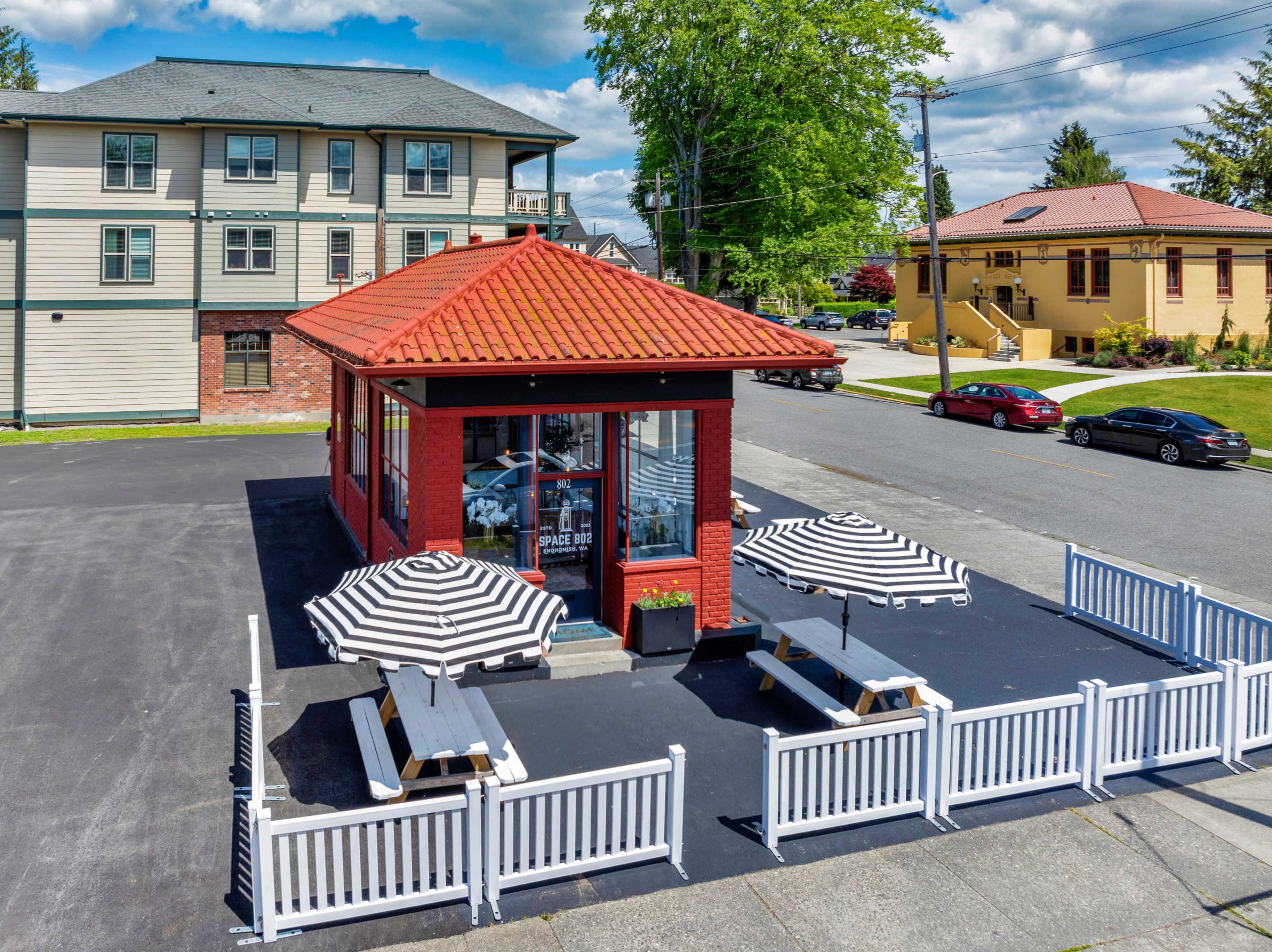 The image shows a red brick building with a tiled roof and outdoor seating, surrounded by a white picket fence and large striped umbrellas.