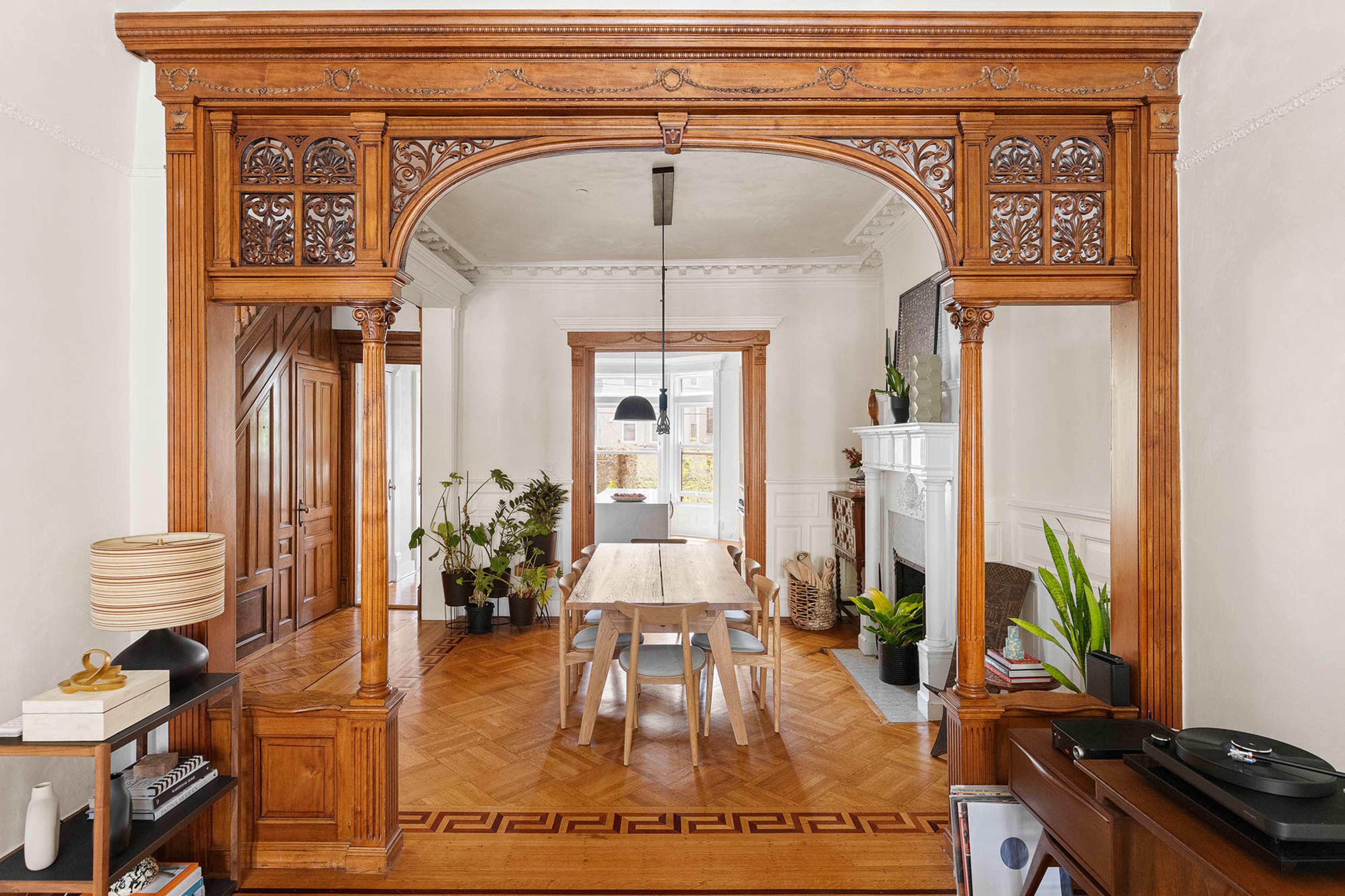 The image features a room with an ornate wooden archway leading to a dining area with a long table surrounded by plants, a pendant light, and a fireplace.