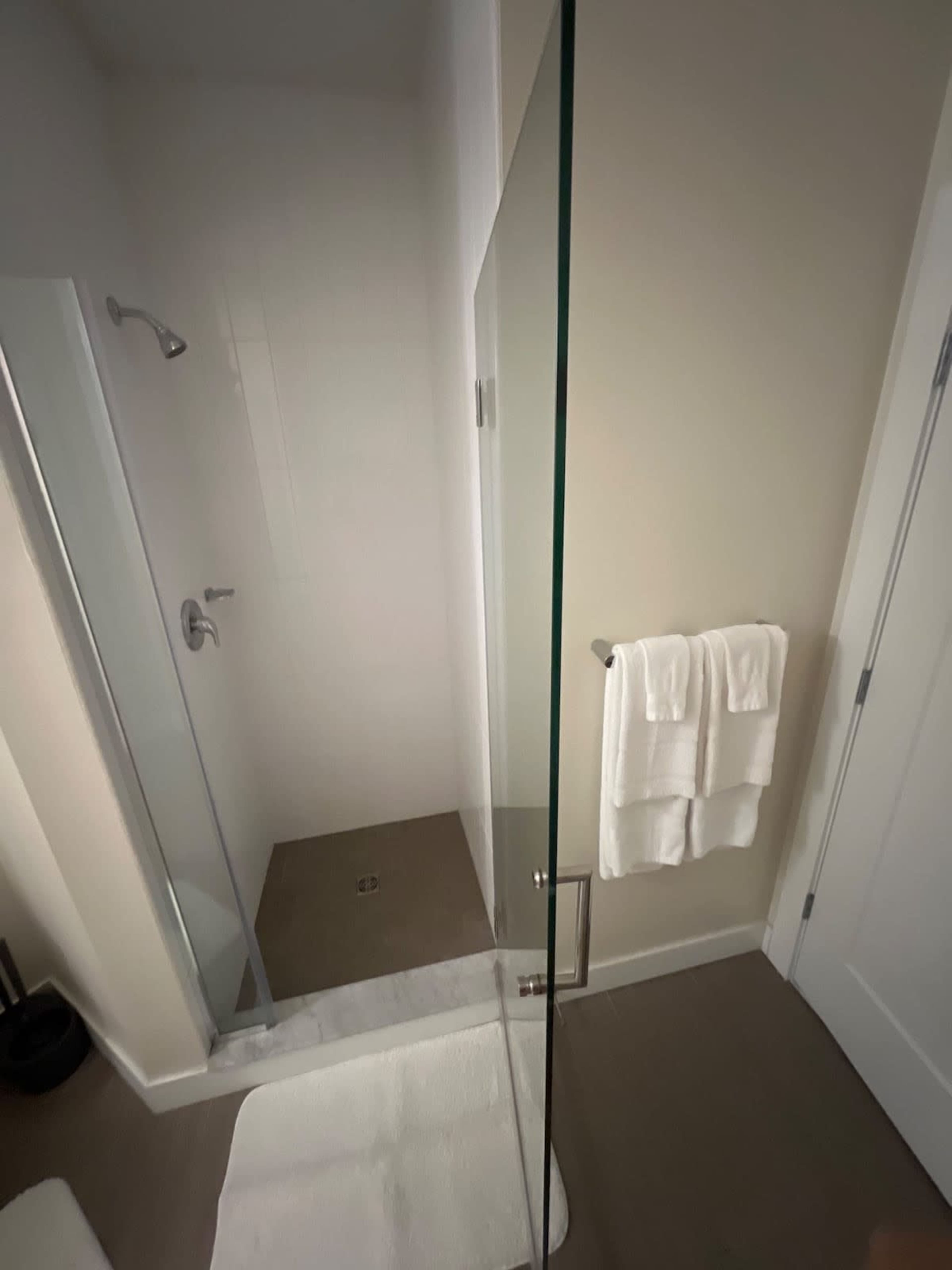 The image shows a modern bathroom with a walk-in shower featuring a glass door, a white bath mat on the floor, and neatly arranged white towels hanging on a rack.