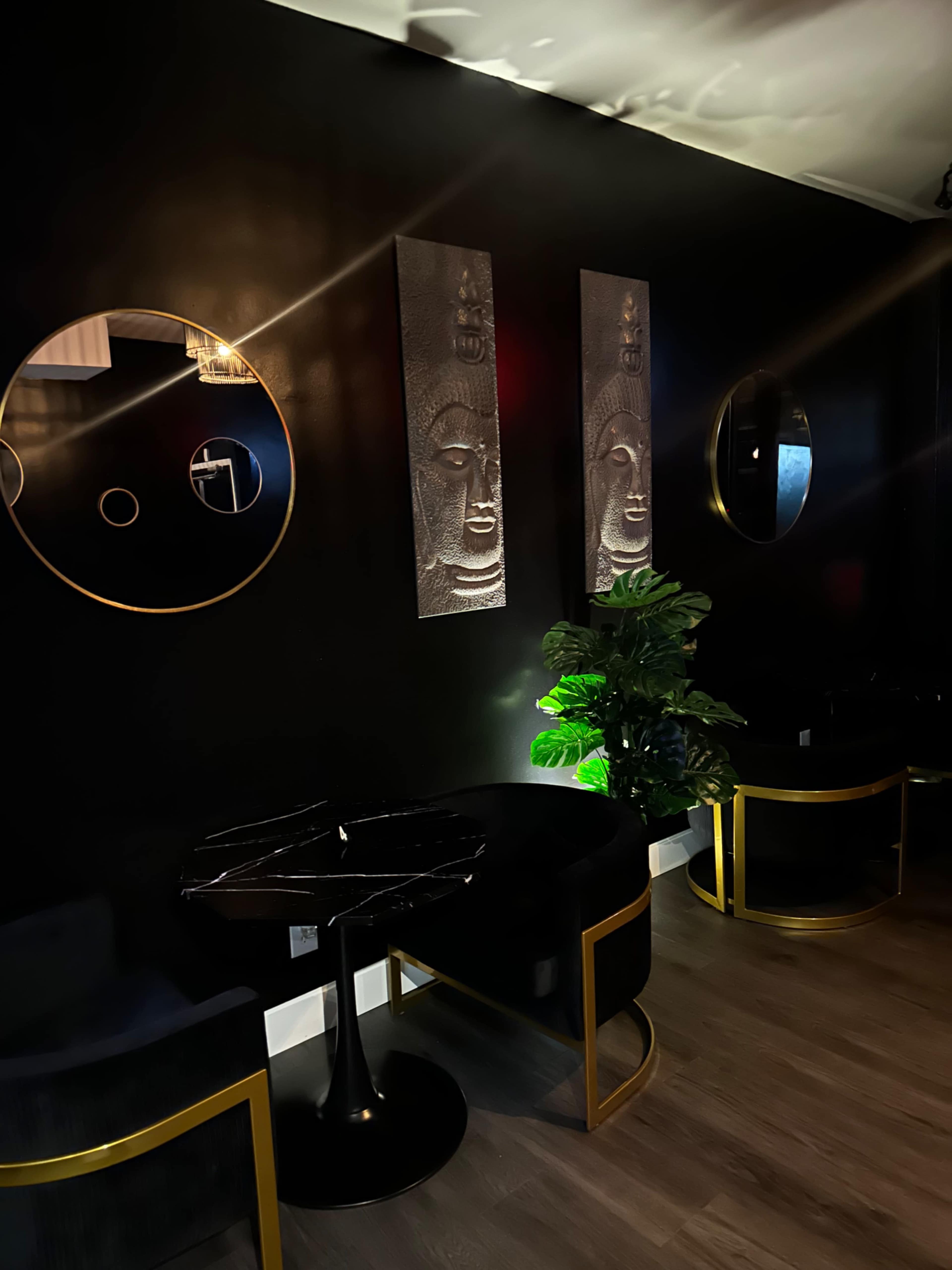 The image shows a dimly lit interior with black walls adorned with two textured wall art pieces of Buddha faces, circular mirrors, and modern furniture featuring gold accents.