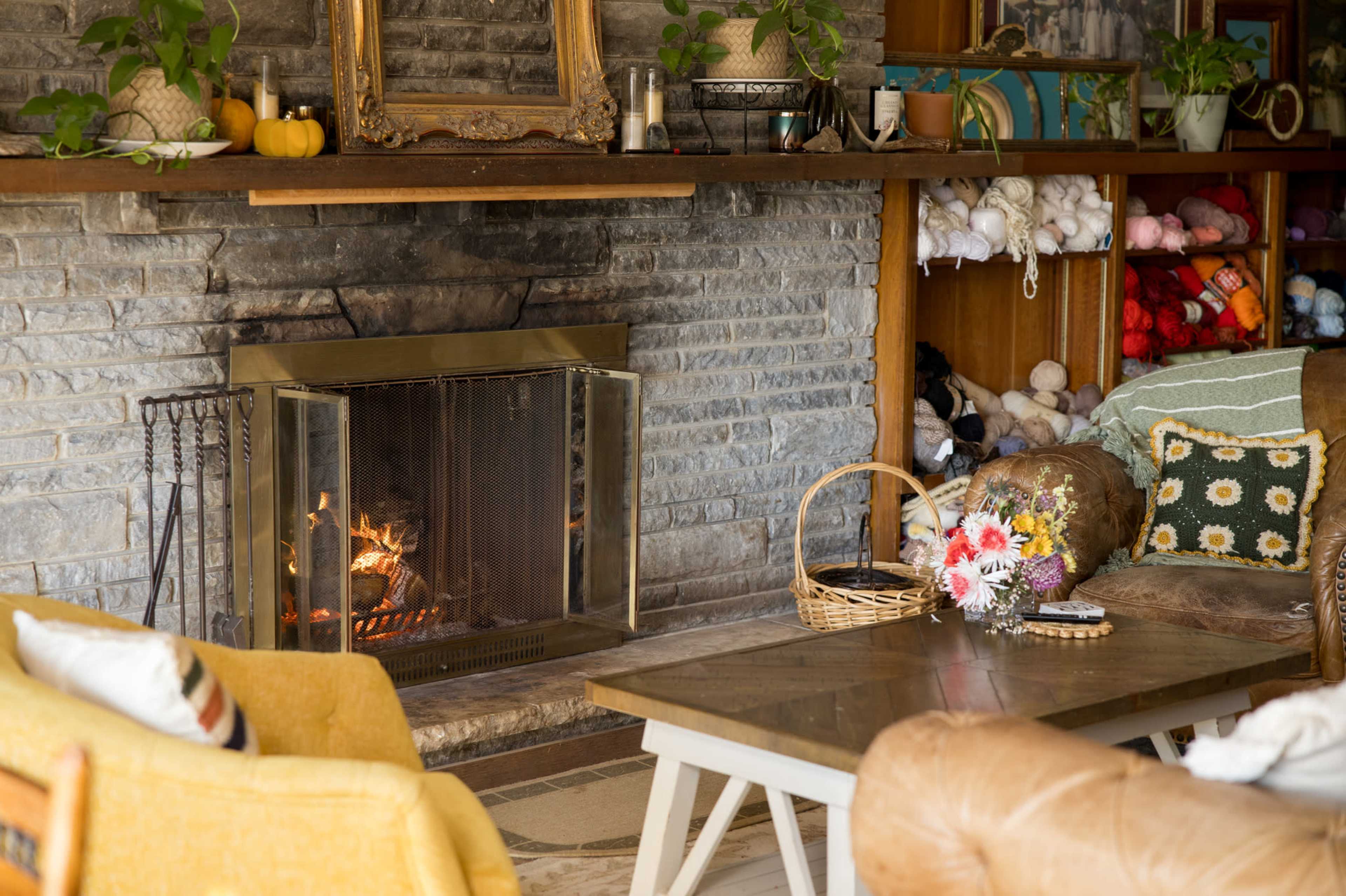 A cozy living room features a stone fireplace with a small fire burning, surrounded by wooden shelves filled with knitting supplies and plants.