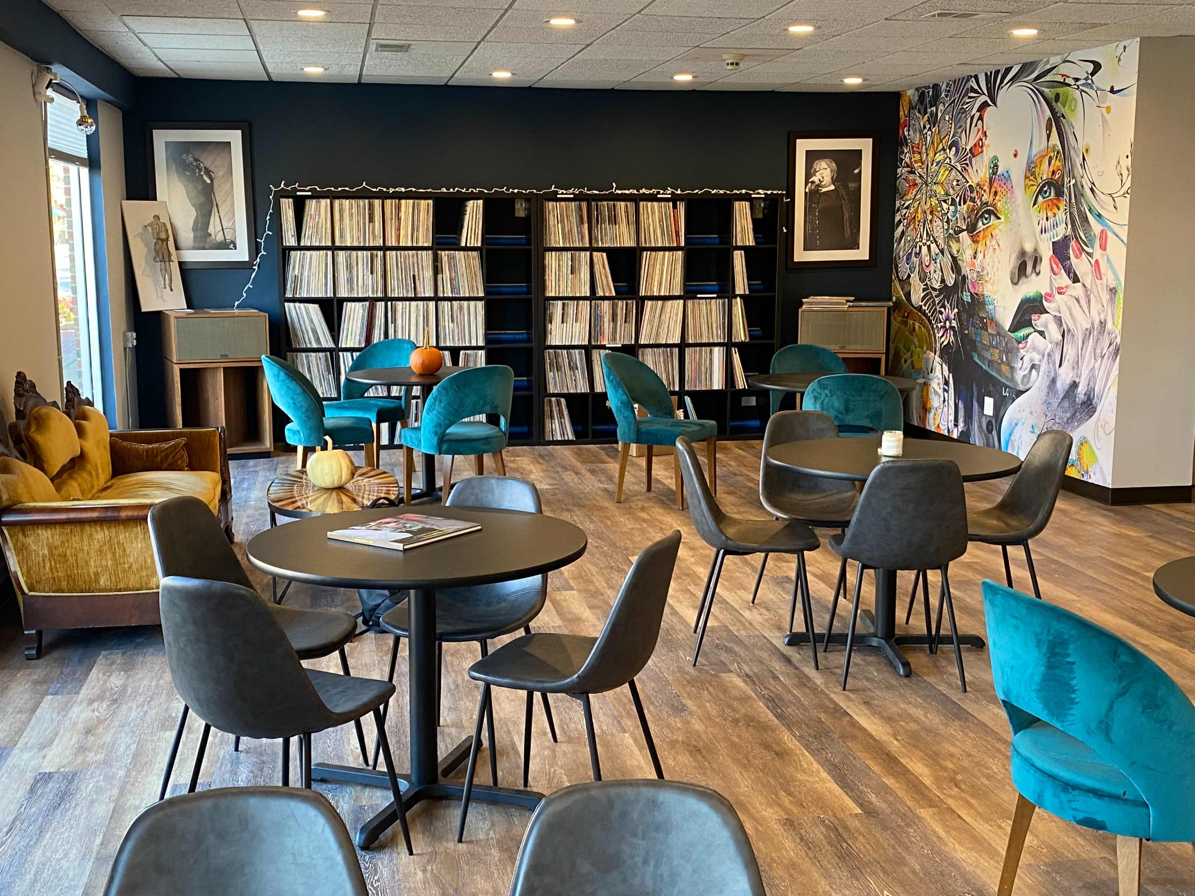 A cozy lounge area featuring tables and chairs, a vinyl record shelf, and a wall adorned with a colorful mural.