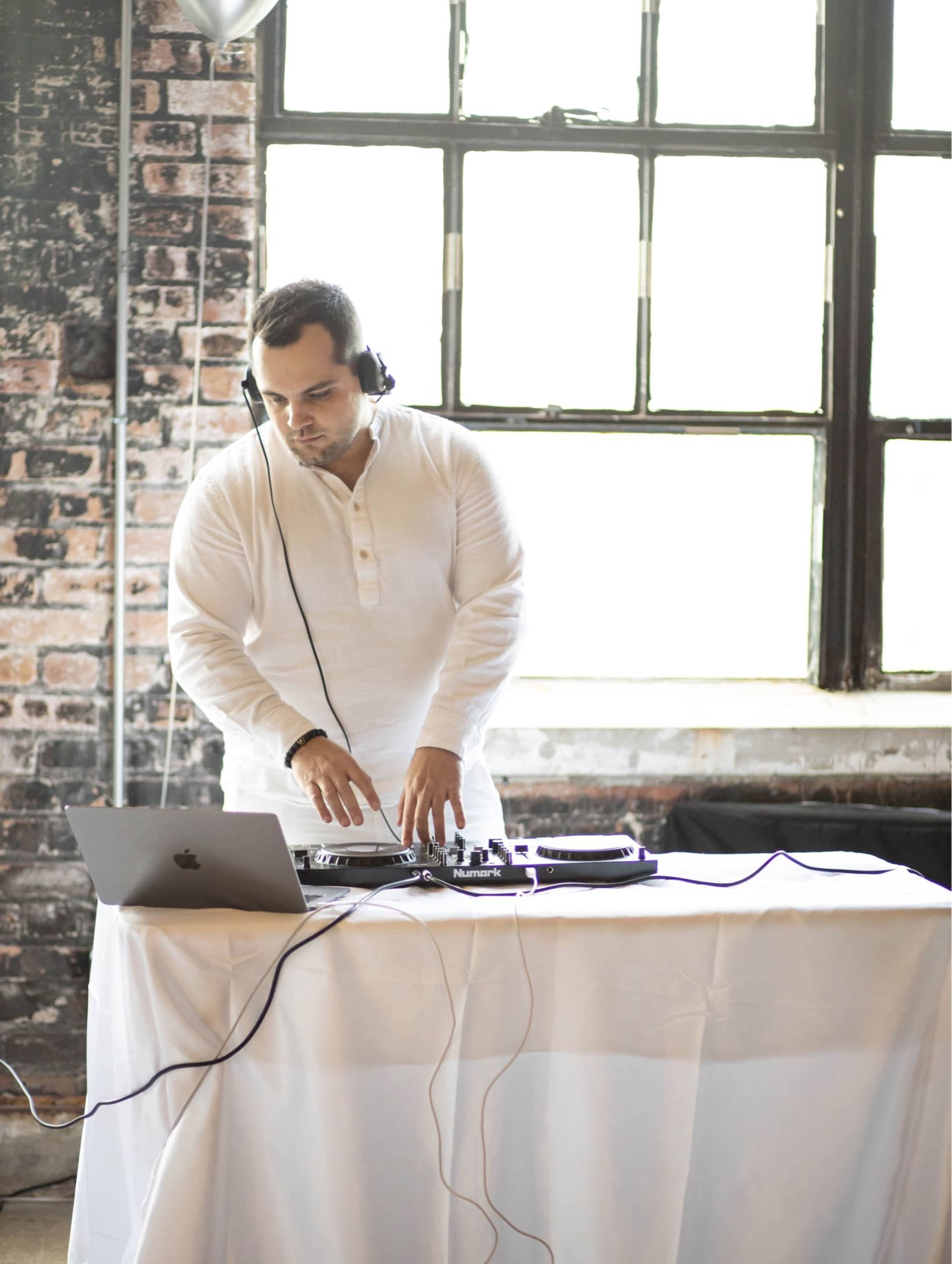 A DJ is performing at a table with a laptop and turntables set up in a room with large windows and exposed brick walls.