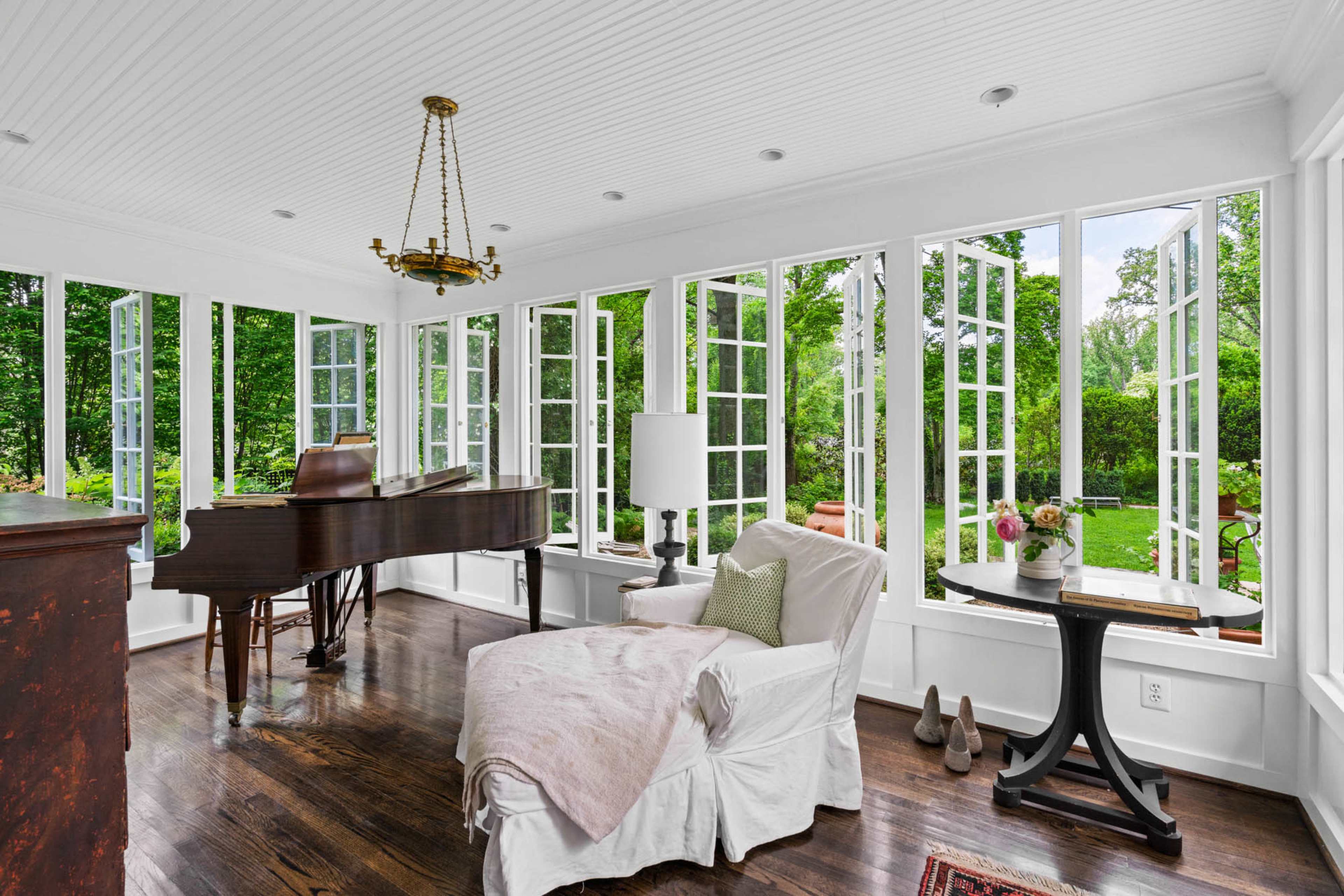 The image depicts a bright, airy sunroom with large windows, a grand piano, a chaise lounge, and a round table with a lamp and decorative items.