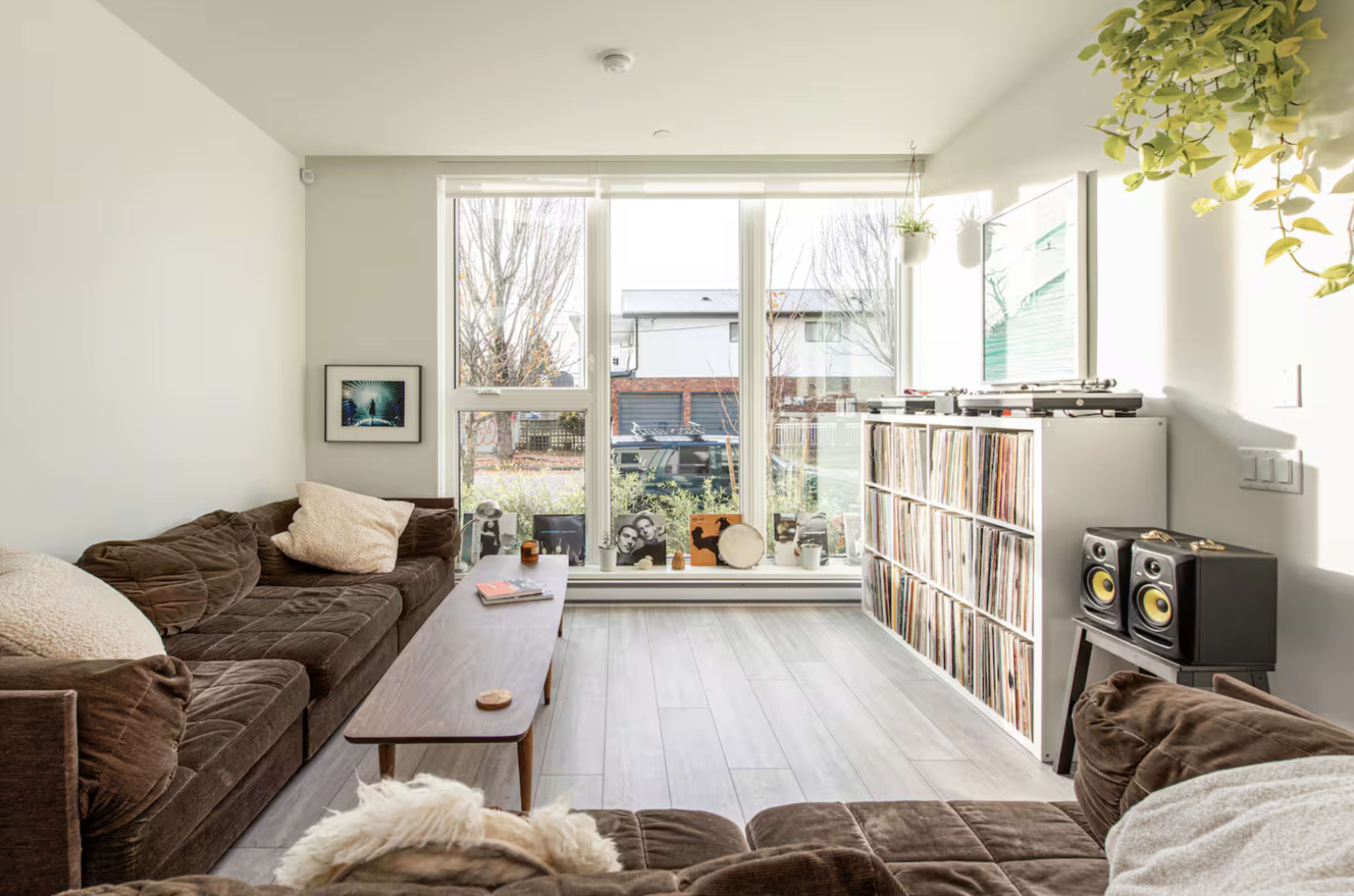 A modern living room features two brown couches, a wooden coffee table, a record shelf with speakers, and large windows that overlook an outdoor area.