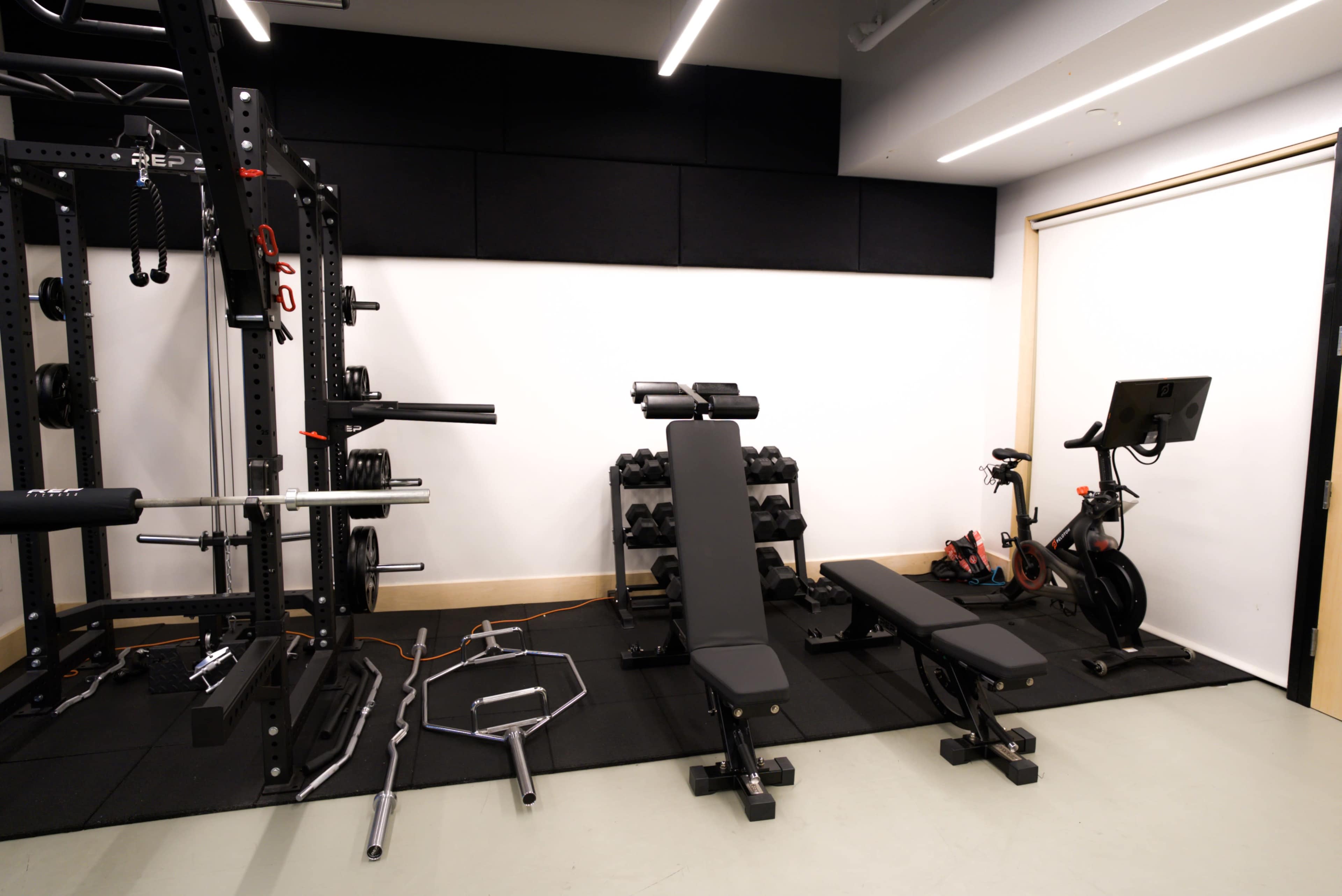 A gym space featuring a power rack, dumbbell weights, two flat benches, a stationary bike, and several barbells arranged on a black mat.