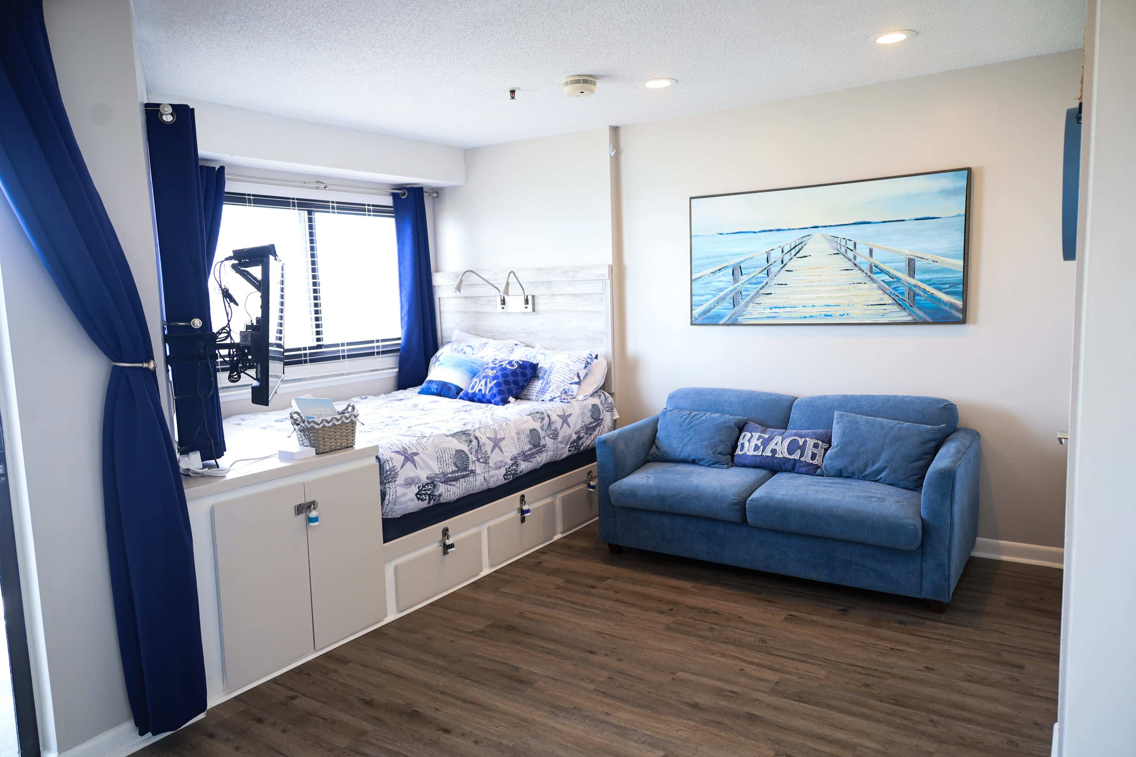 A small bedroom featuring a bed with blue and white bedding, a blue sofa with "BEACH" decor, and a large painting of a pier on the wall.