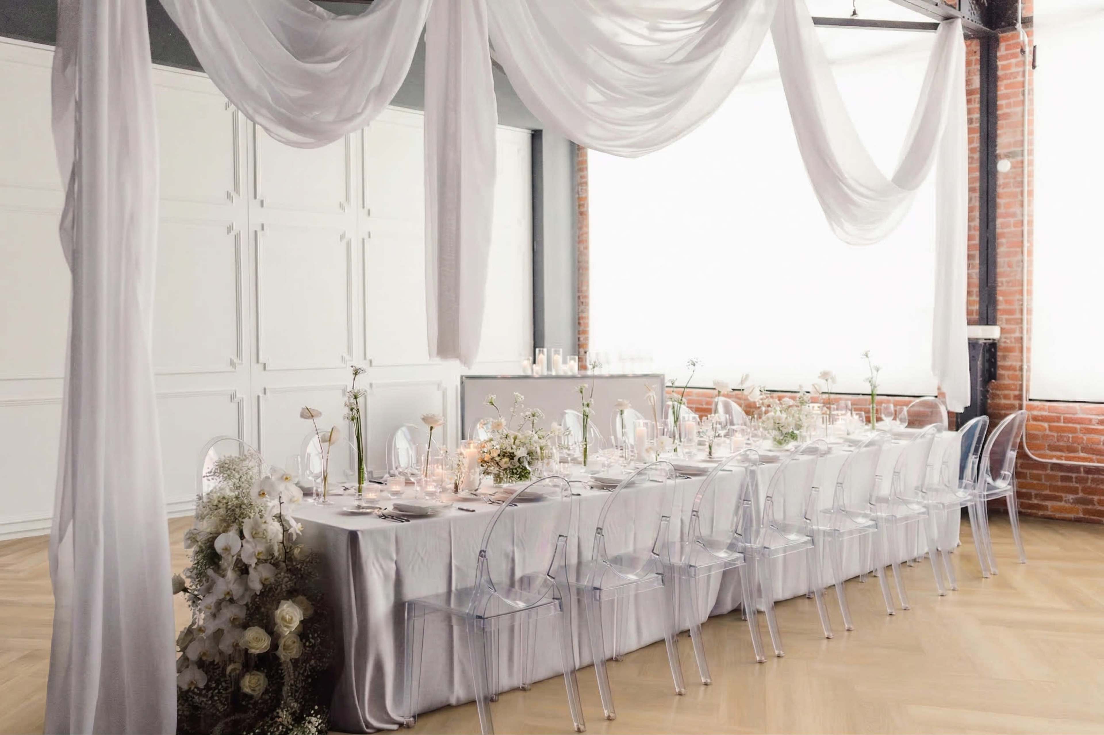 The image shows a long dining table elegantly set with white tableware and surrounded by clear acrylic chairs in a decorated room with flowing white fabric draped from the ceiling.