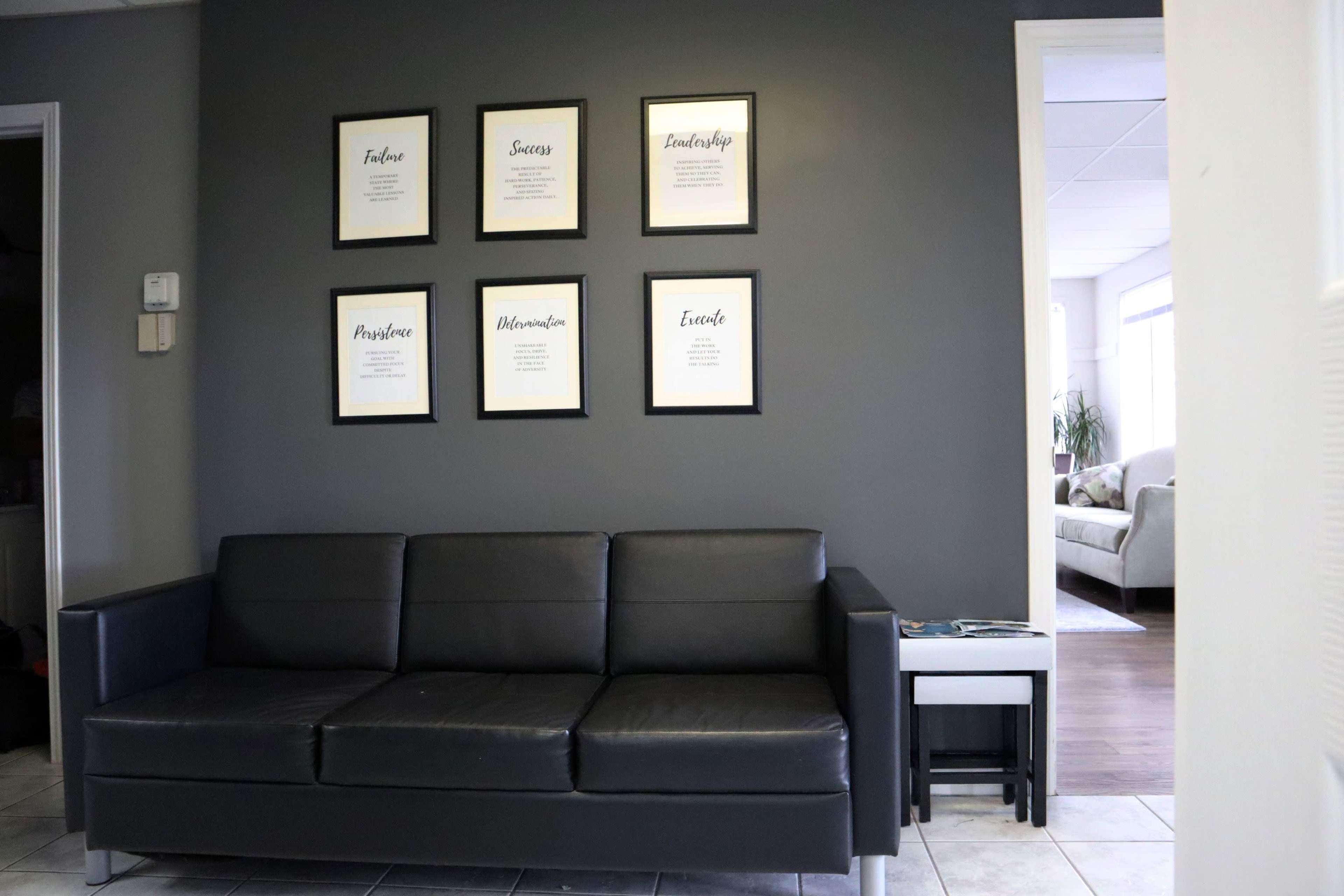 A black sofa is positioned against a gray wall adorned with framed motivational quotes, with a glimpse of a well-lit room visible in the background.