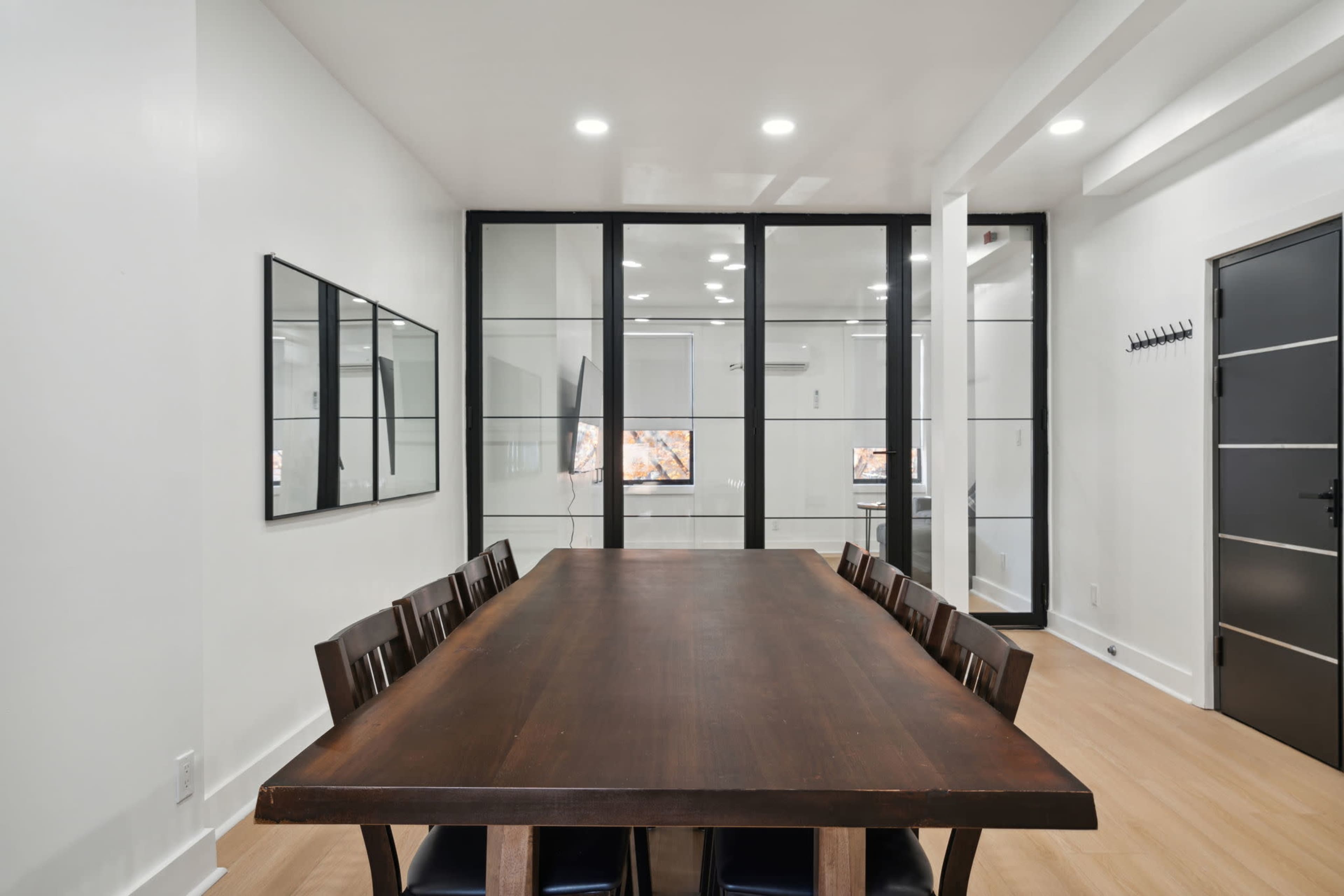 A long wooden dining table with eight chairs is set in a bright room featuring glass walls and a large mirror.