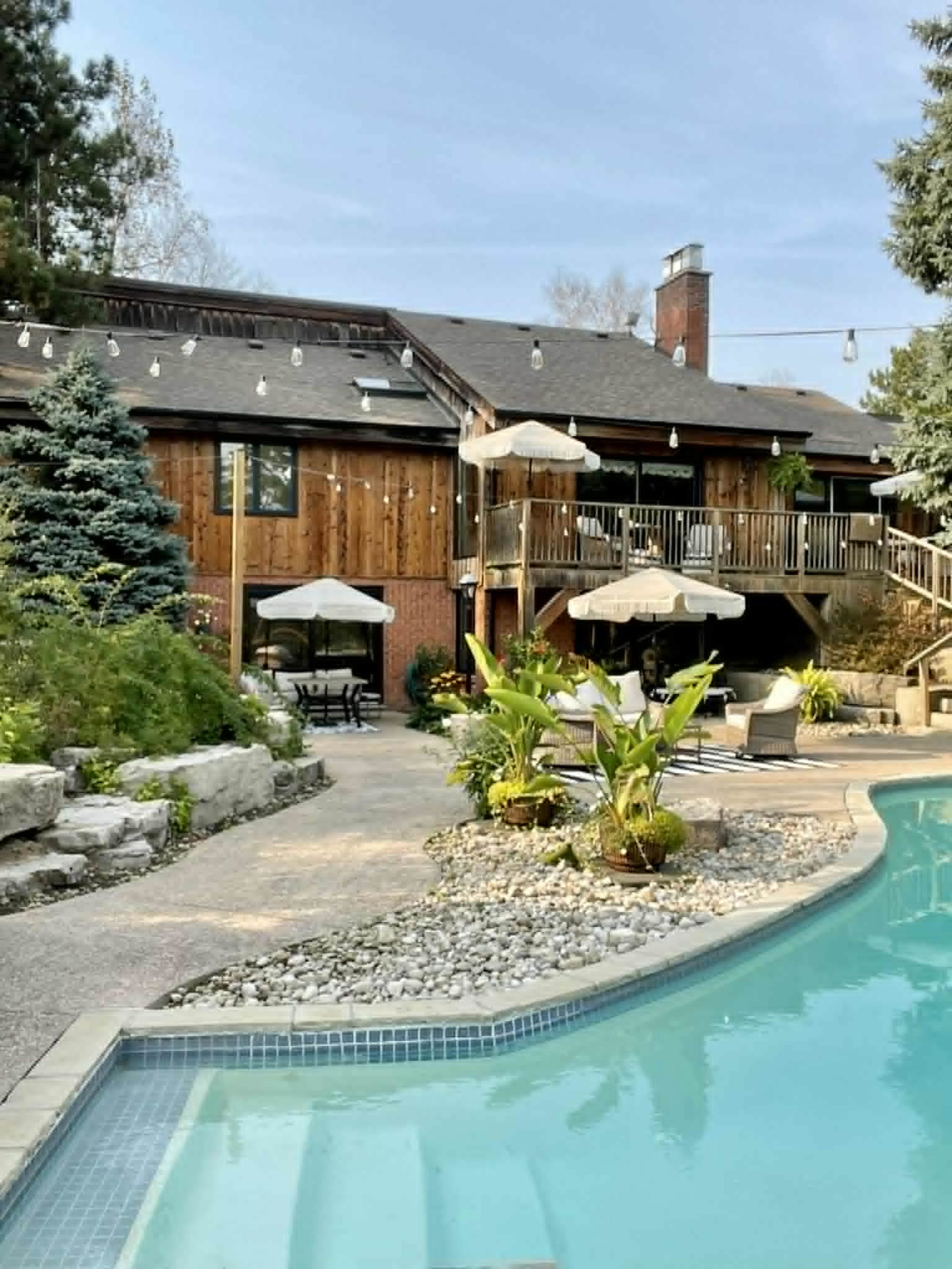 The image depicts a backyard with a swimming pool in the foreground and a wooden house with a deck and patio area in the background, surrounded by landscaped plants and trees.