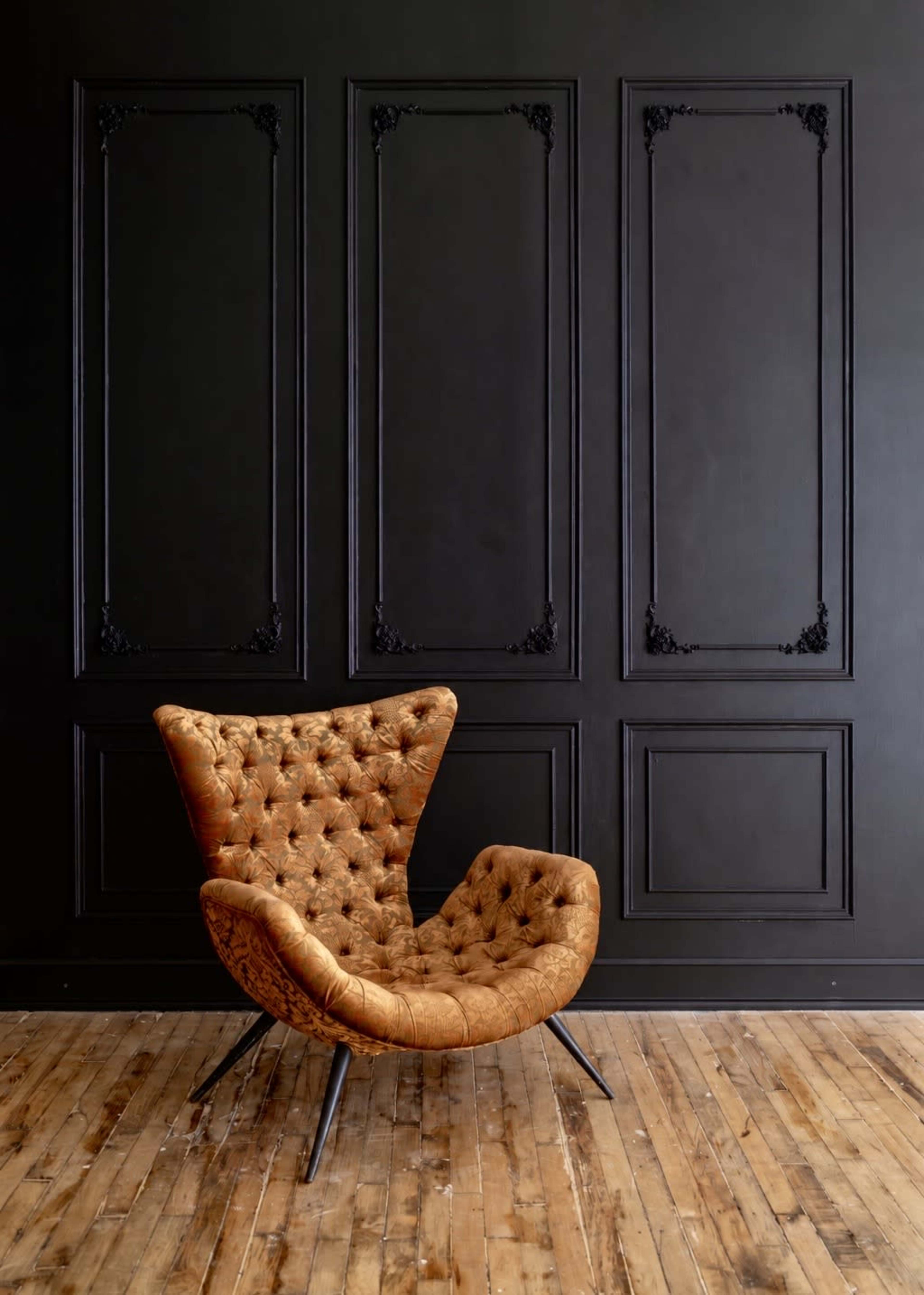 A tufted orange chair is placed against a black paneled wall with intricate moldings, on a wooden floor.