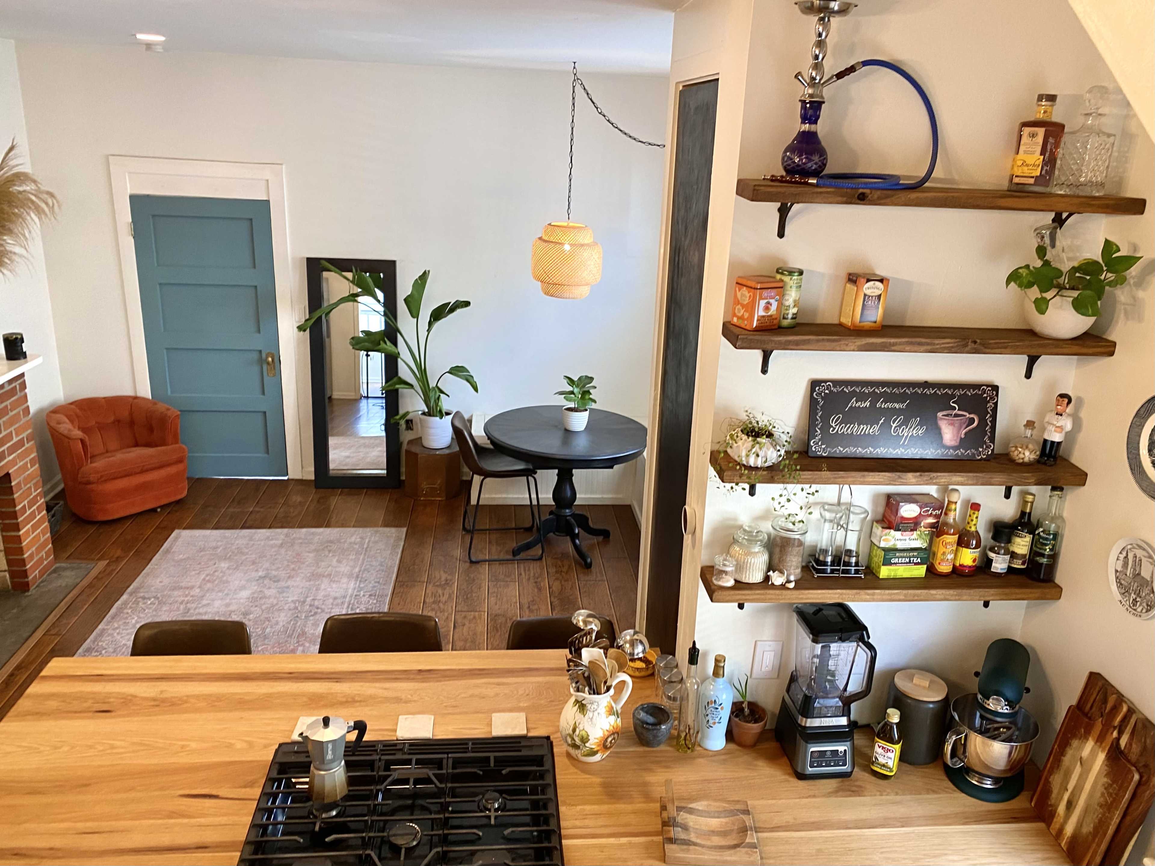 The image shows an open-concept kitchen and living area with a wooden countertop, shelves displaying various kitchen items, and a cozy seating area featuring an orange chair.
