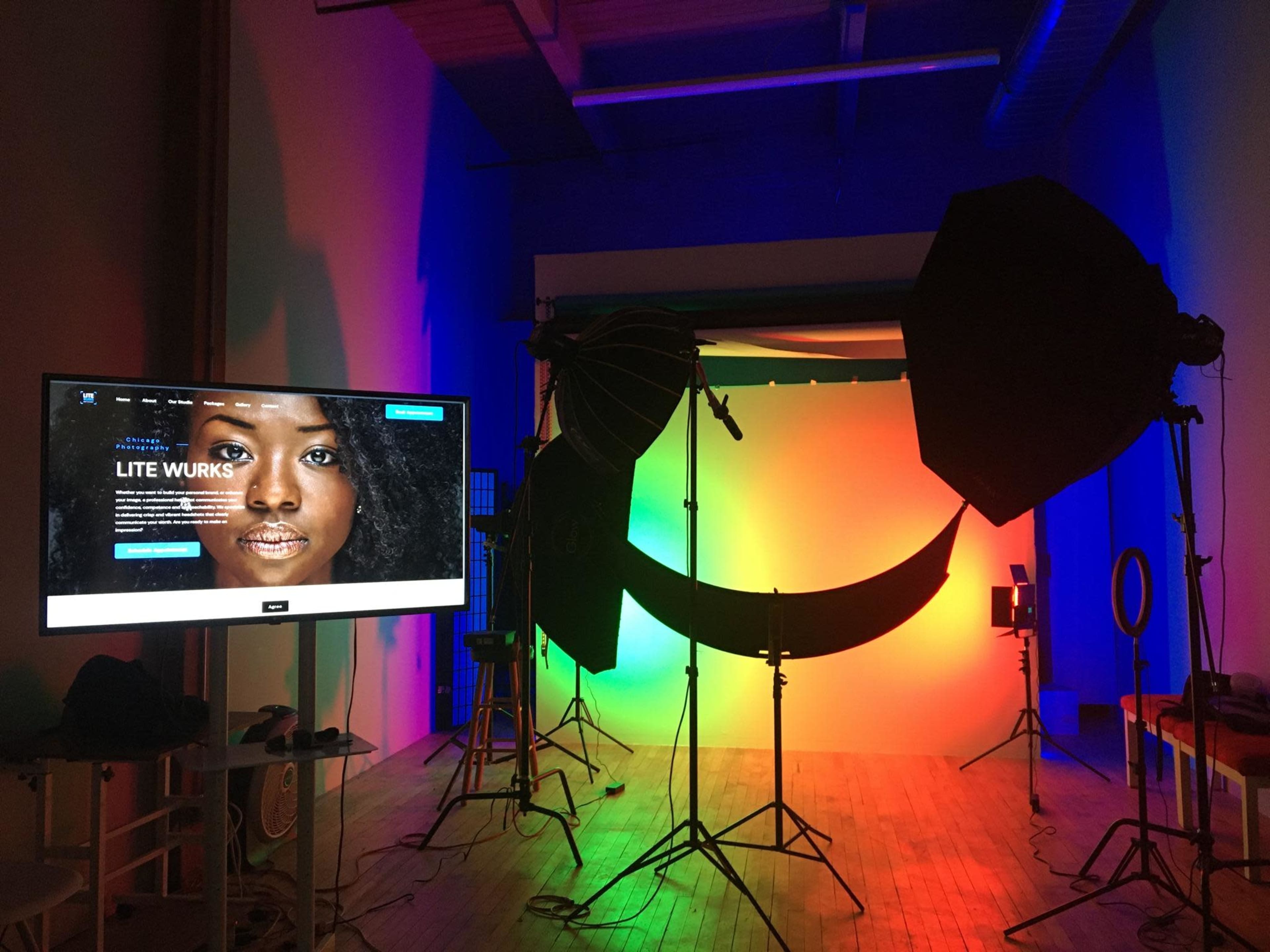 A photography studio setup with colorful lighting, a backdrop, and a monitor displaying a website.