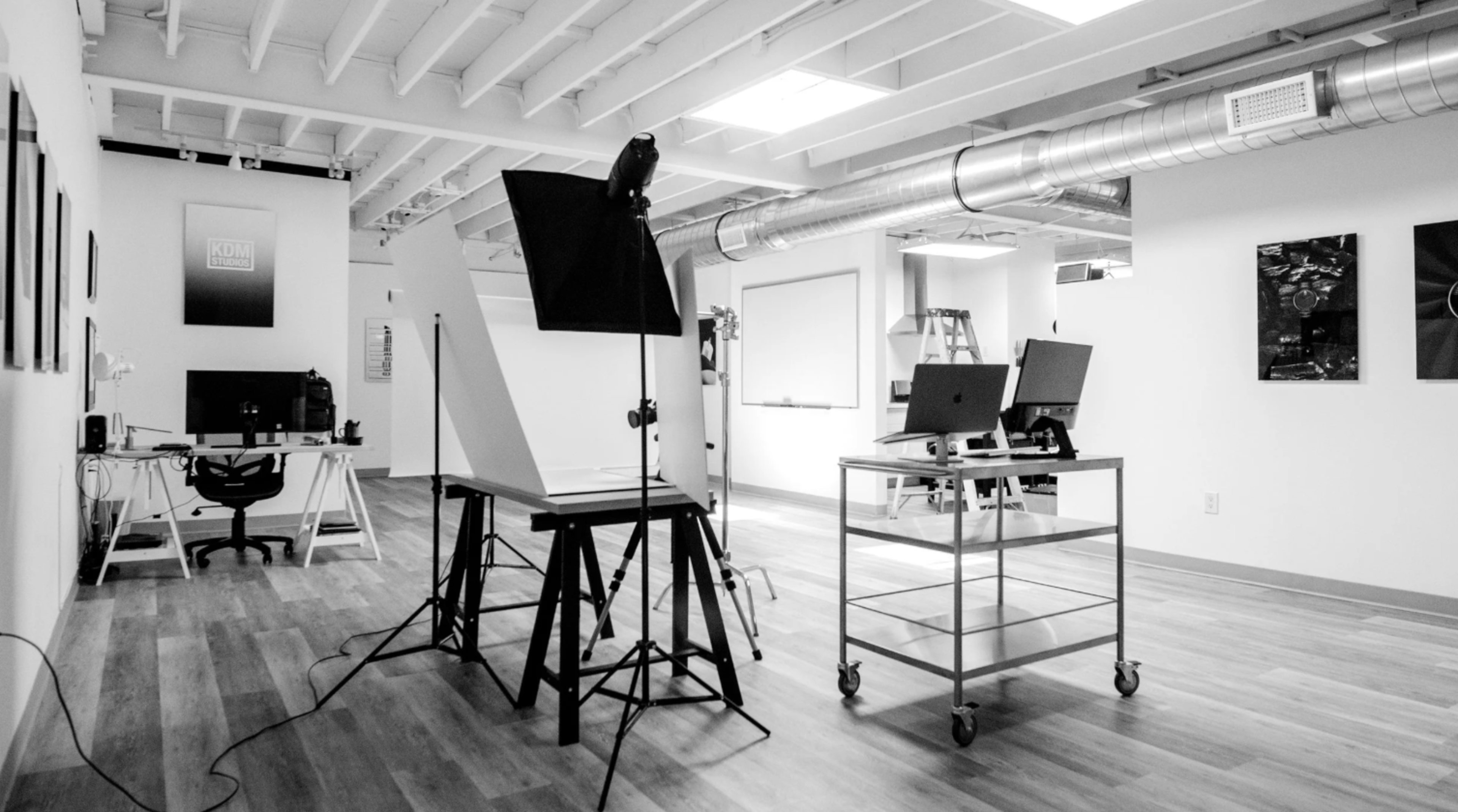 The image shows a spacious, well-lit office area featuring multiple workstations, a photo setup with a light stand, and industrial-style ventilation.
