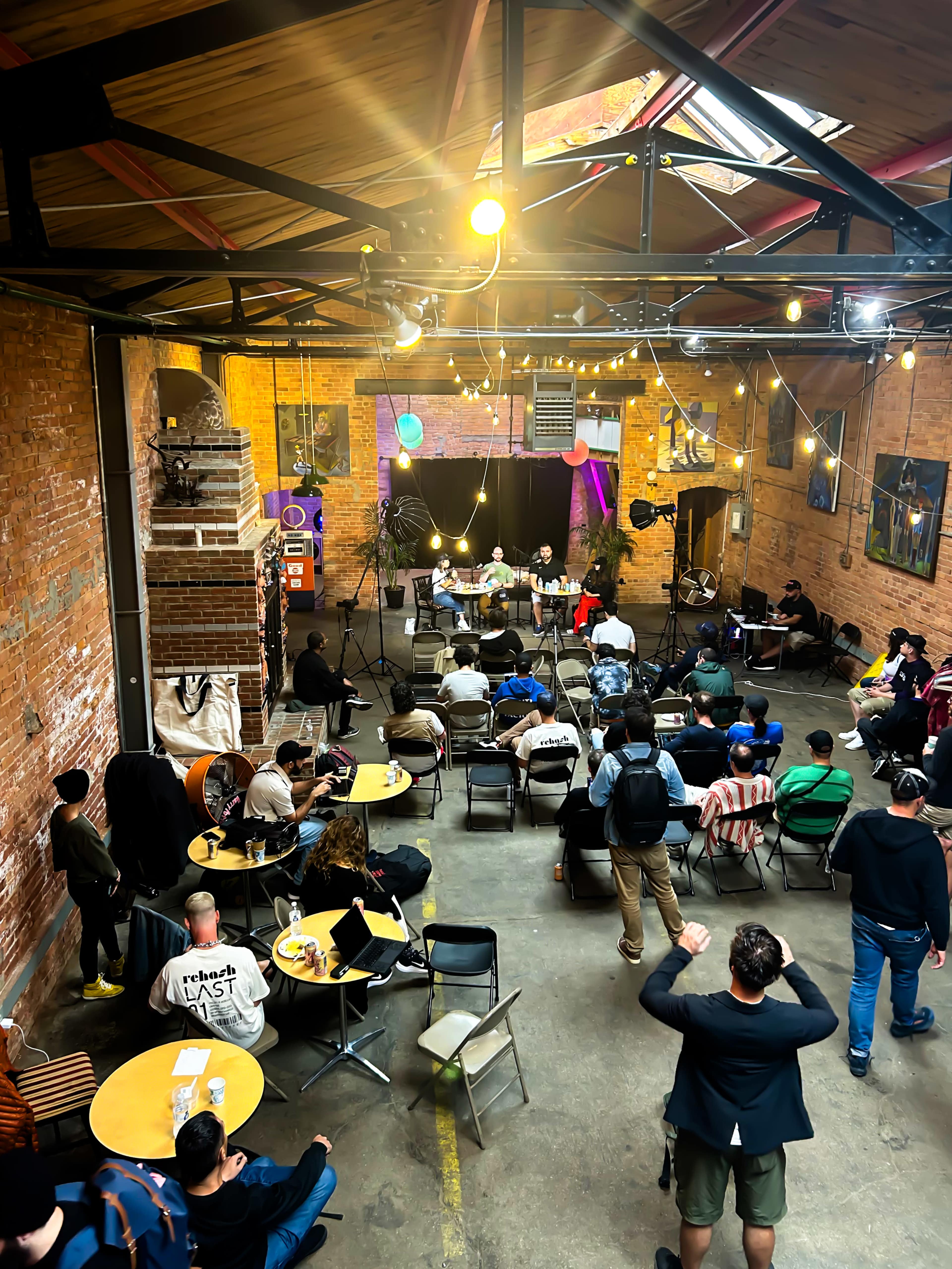 A large crowd sits in a warehouse-style space, facing a stage with colorful lighting as a speaker addresses the audience.