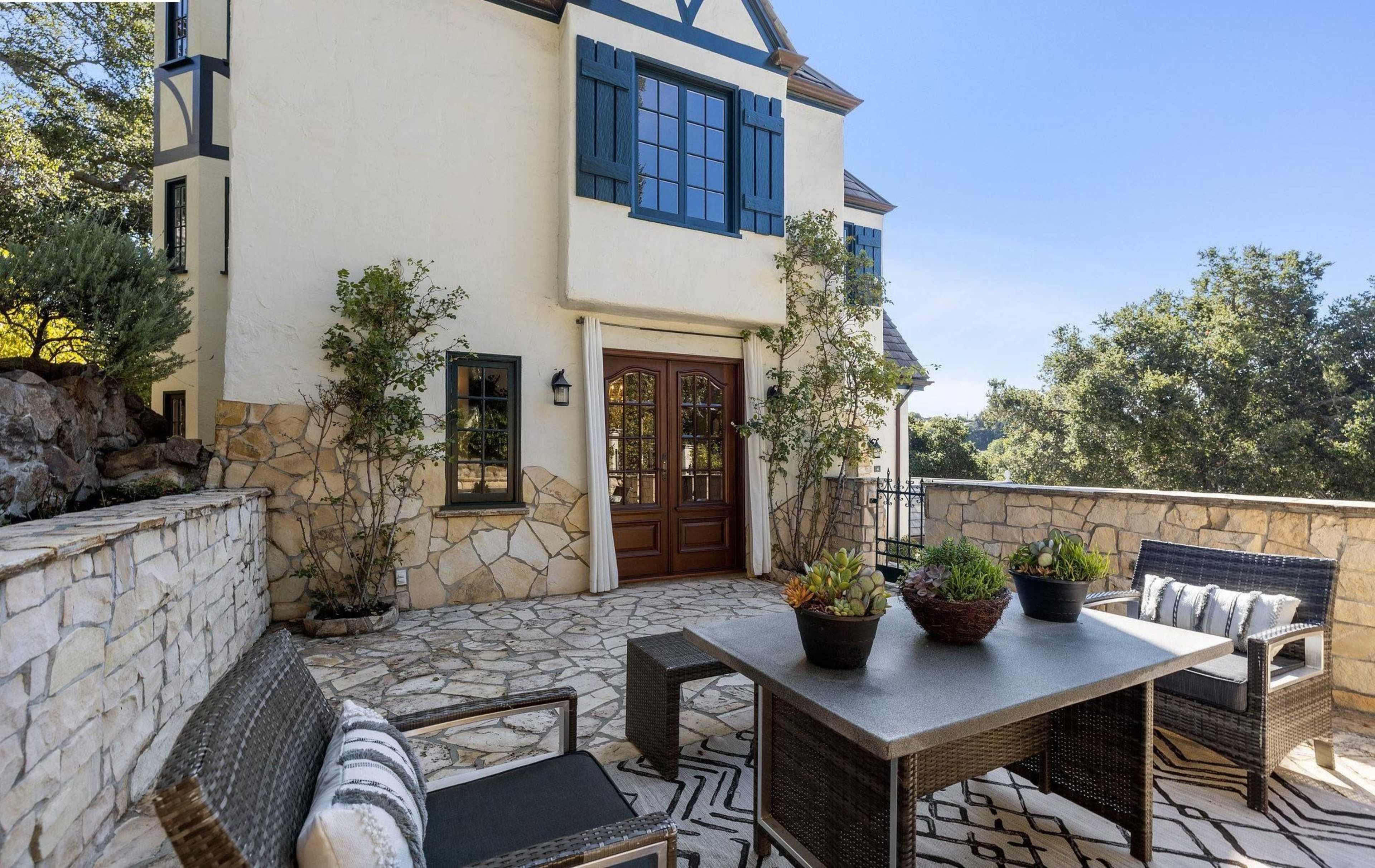 A stone patio features a table and chairs surrounded by potted plants in front of a two-story house with blue shutters.