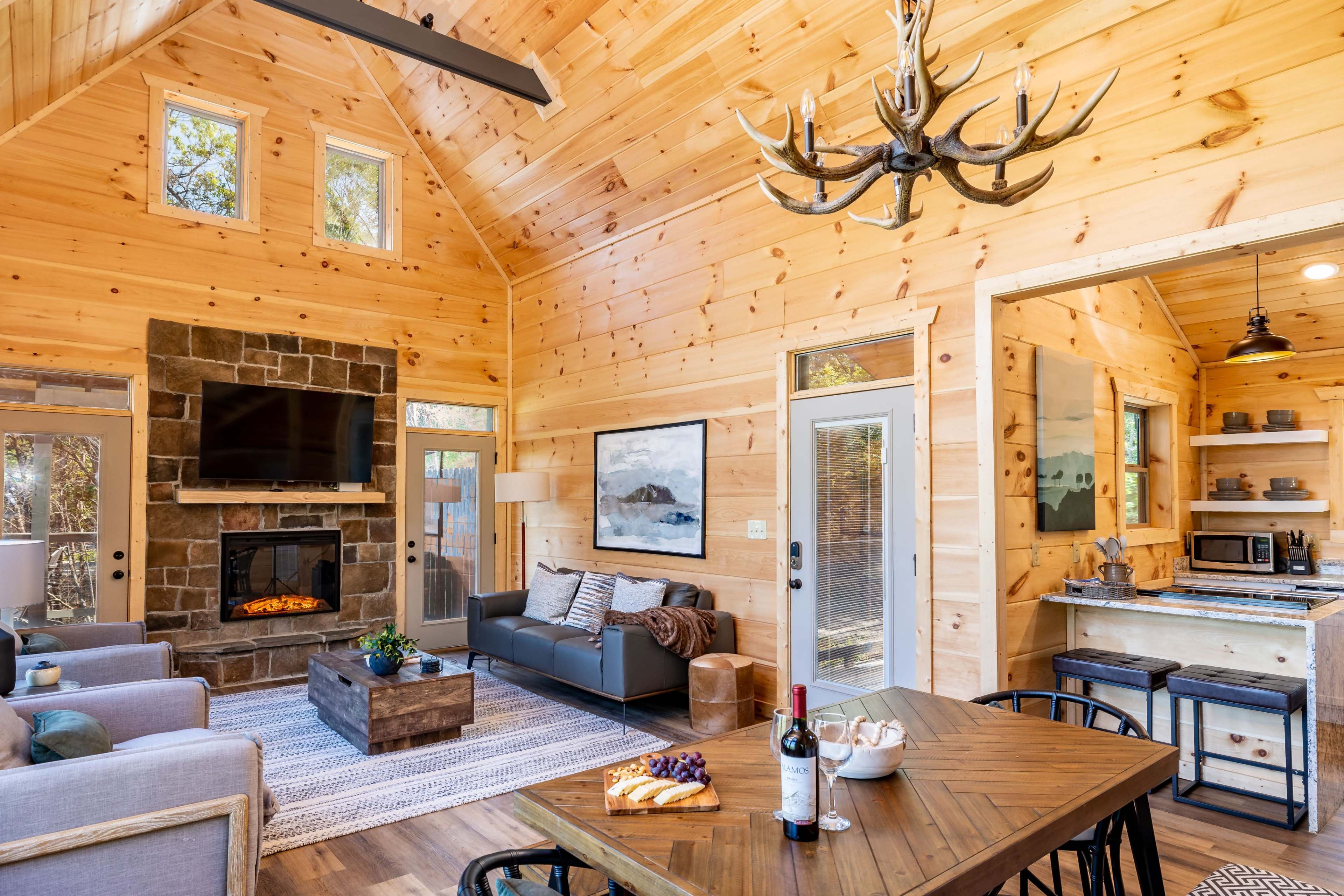 The image shows a cozy wooden cabin interior with a stone fireplace, a television mounted on the wall, a dining table, and a kitchen area visible in the background.