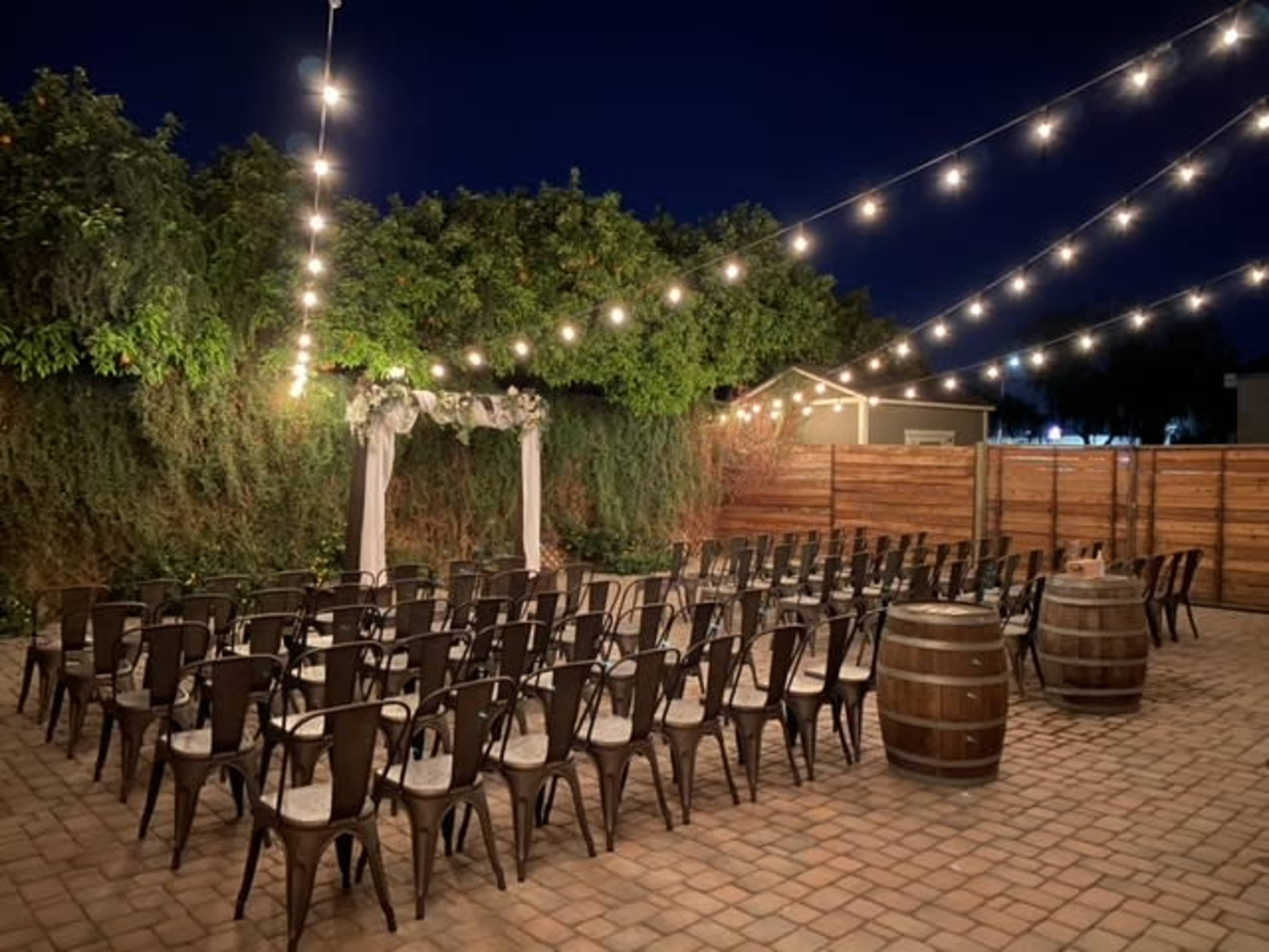 An outdoor event space at night, featuring rows of chairs arranged facing an archway, with string lights overhead and wooden barrels nearby.