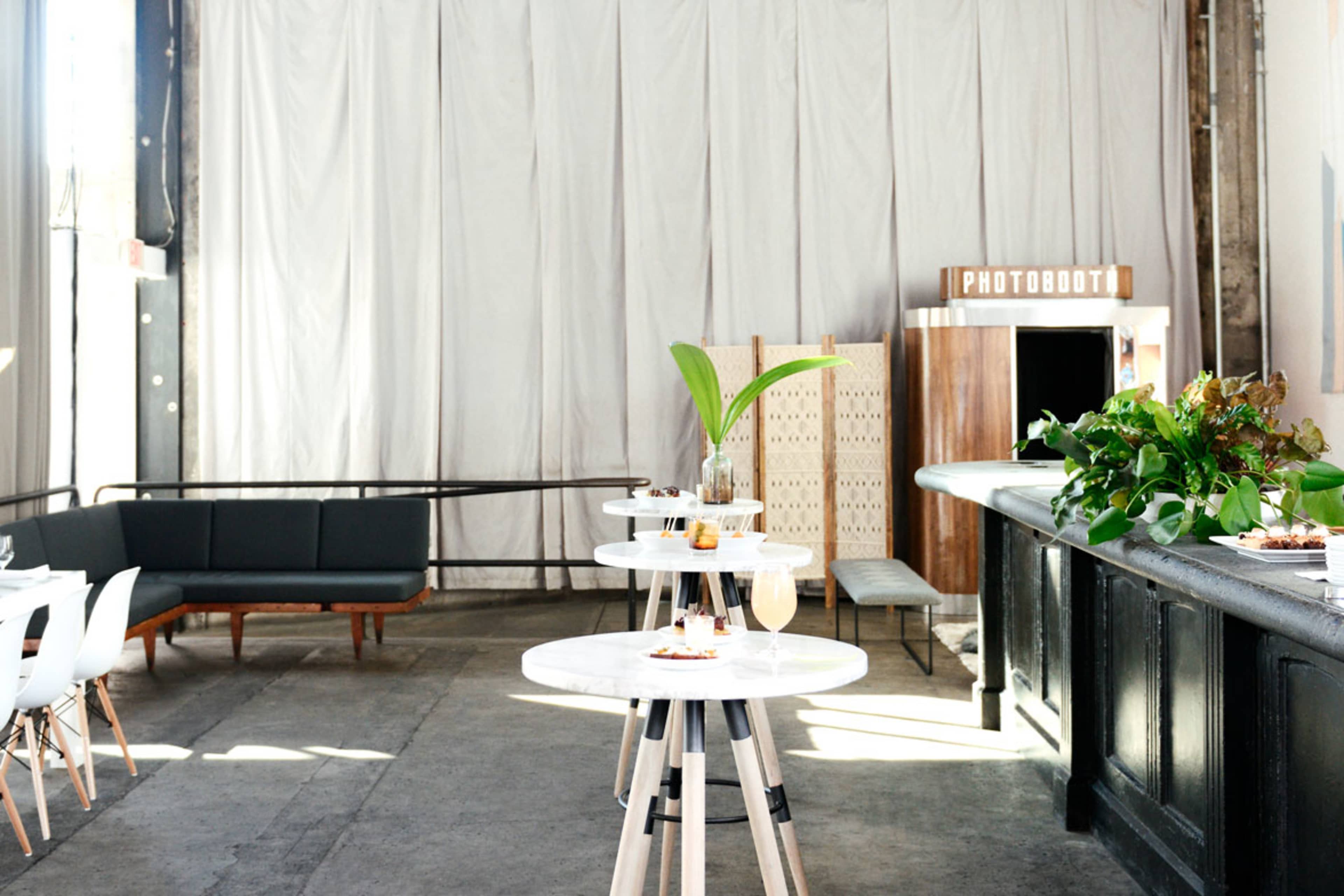 A spacious interior with modern furniture, including tables and a couch, alongside a photobooth and a greenery arrangement.
