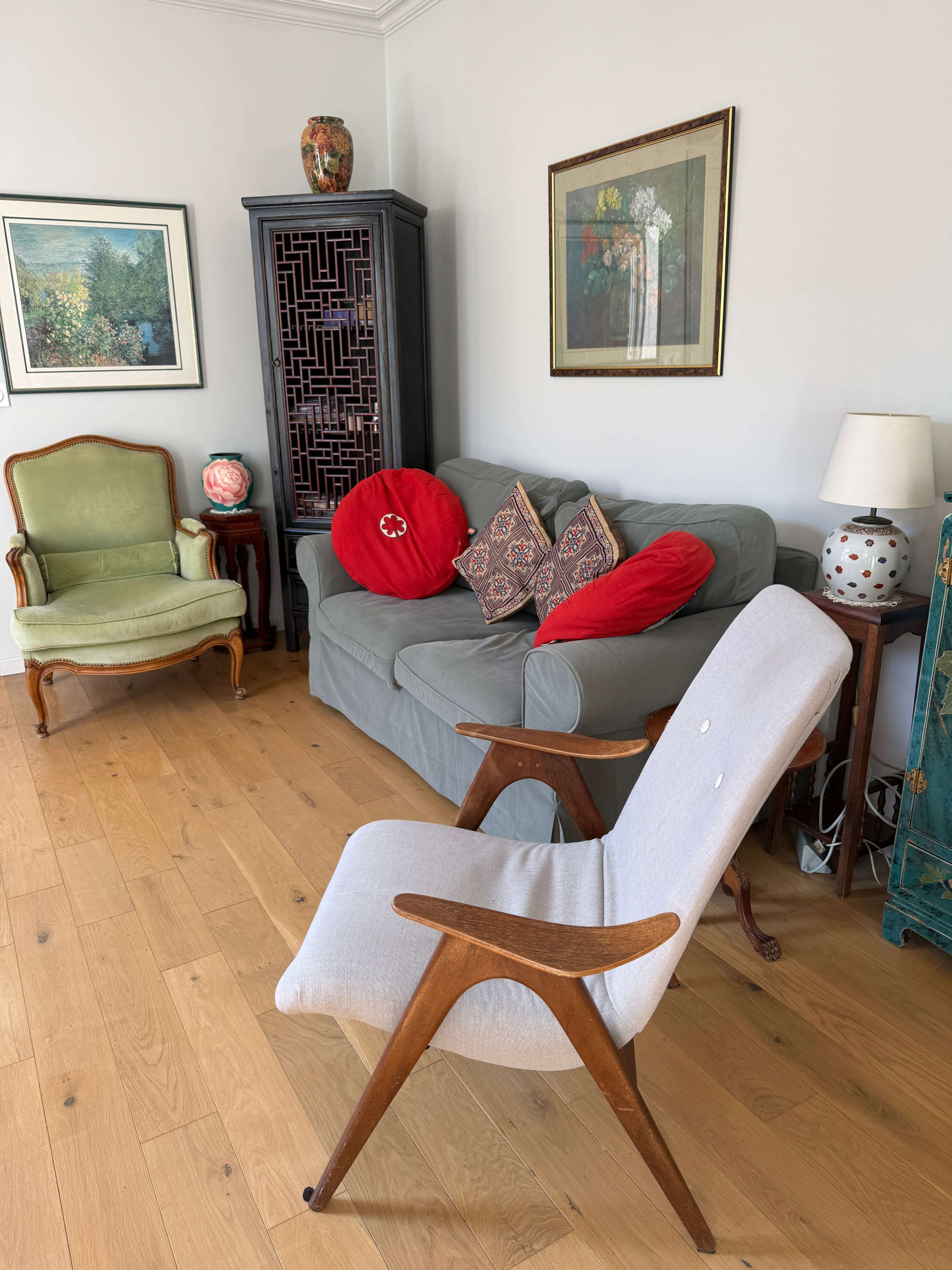 The image depicts a living room with a gray sofa, accented by red pillows, a wooden chair, and various framed artworks on the walls.