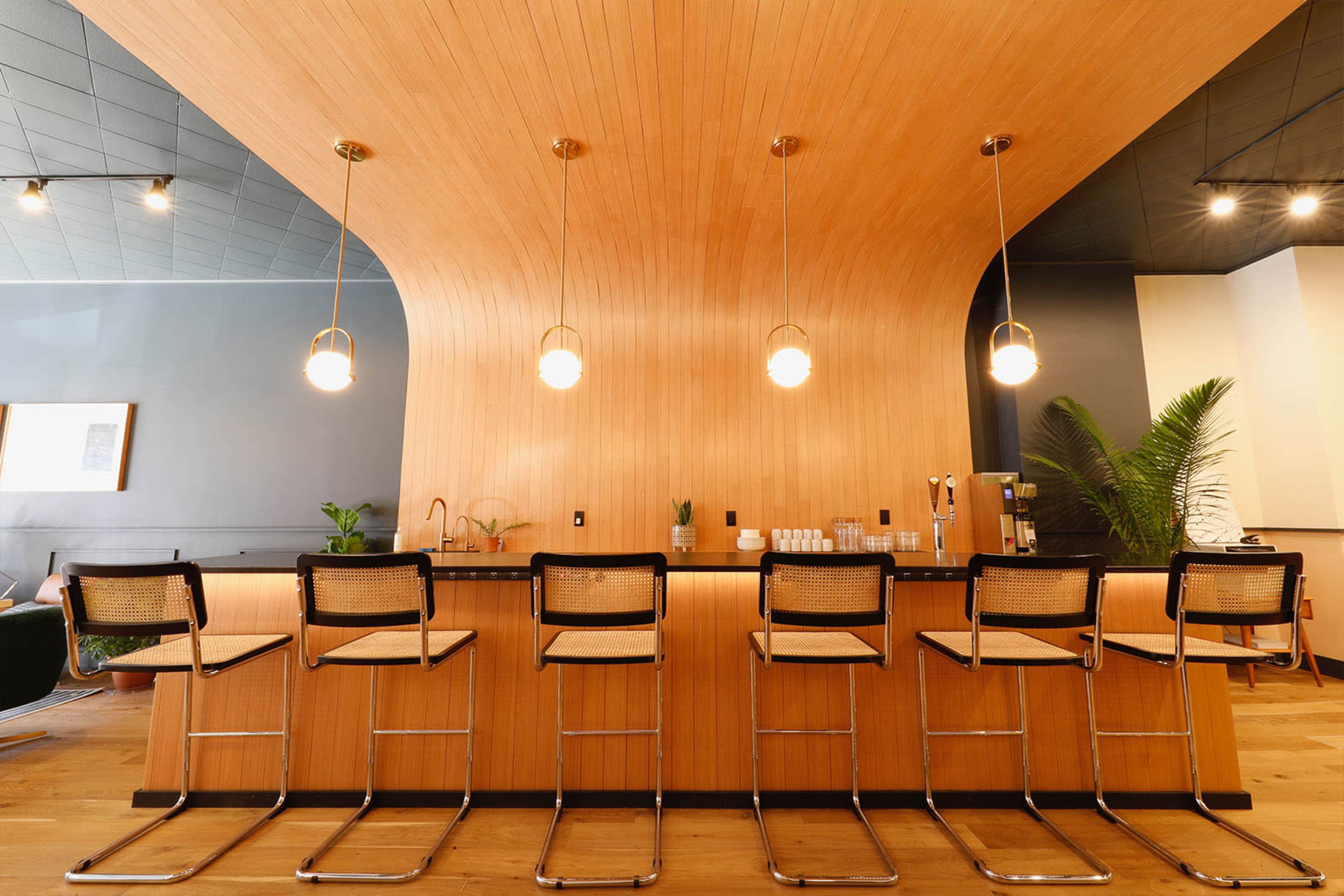 A bar area features a curved wooden ceiling above sleek black stools arranged in front of a countertop with pendant lights.