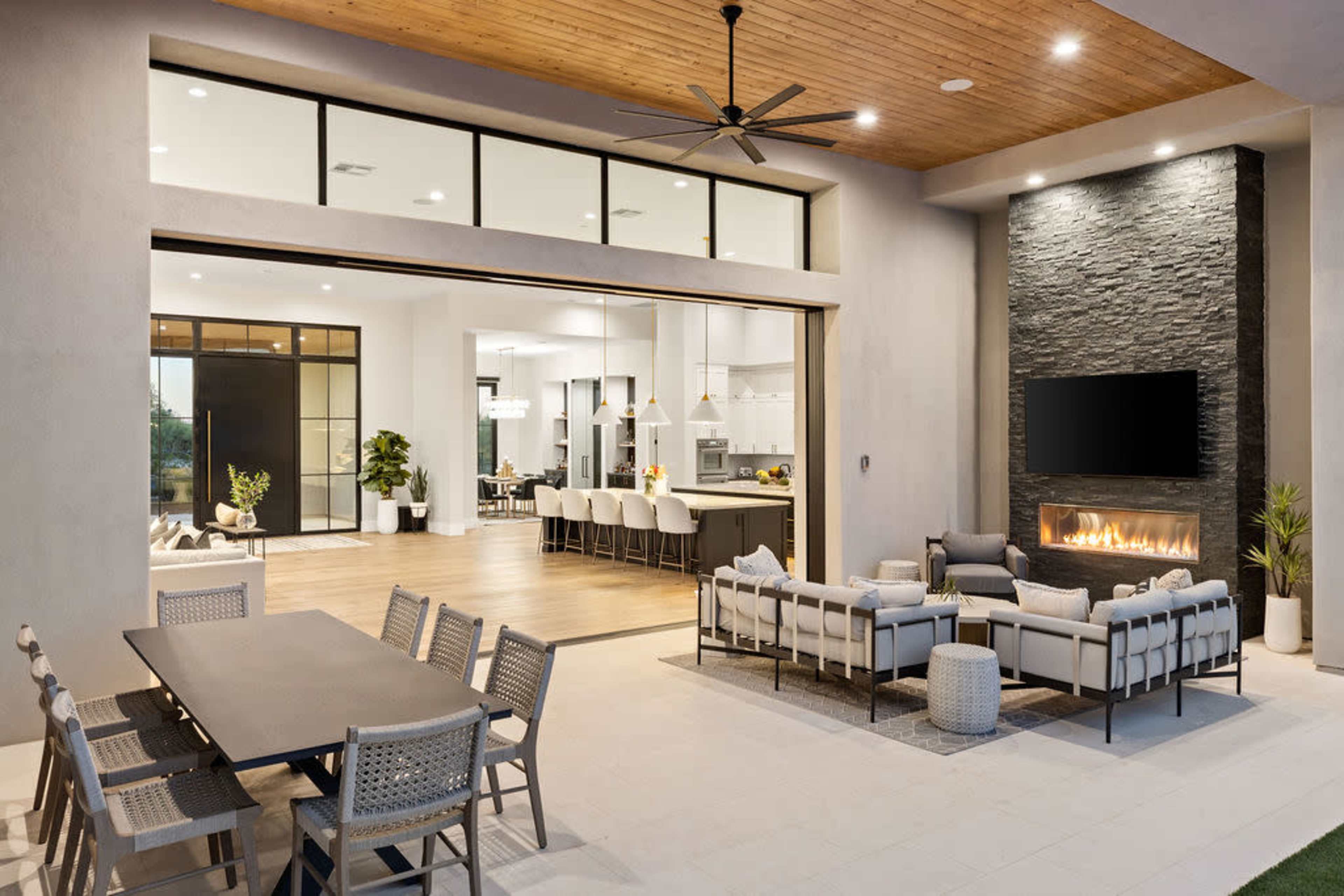 A modern outdoor living space with a seating area and a dining table, adjacent to an open kitchen with large windows and a stone fireplace.