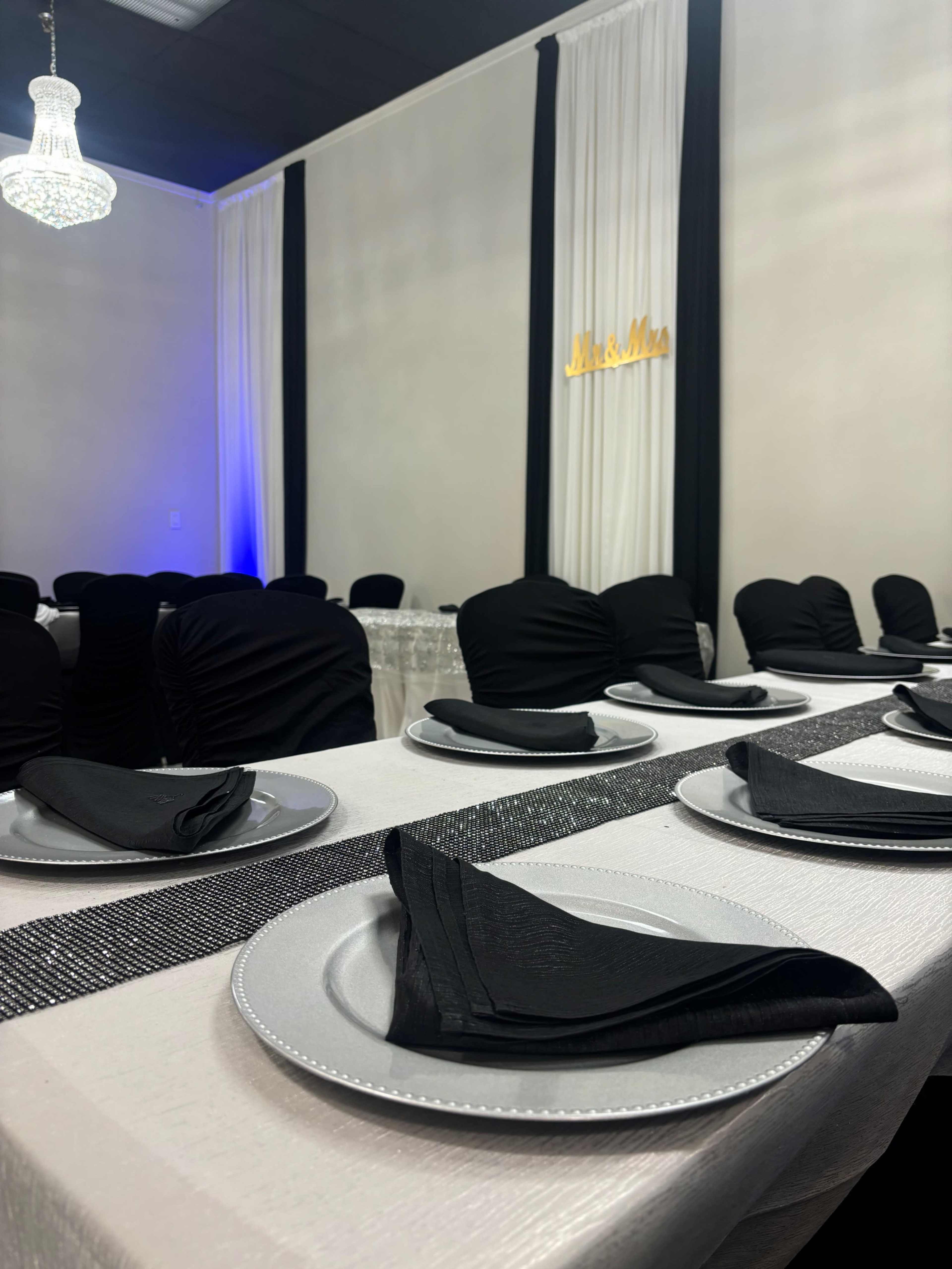 A elegantly set dining table features black napkins and plates against a backdrop of black and white drapes, with a decorative "Mr. & Mrs." sign illuminated nearby.