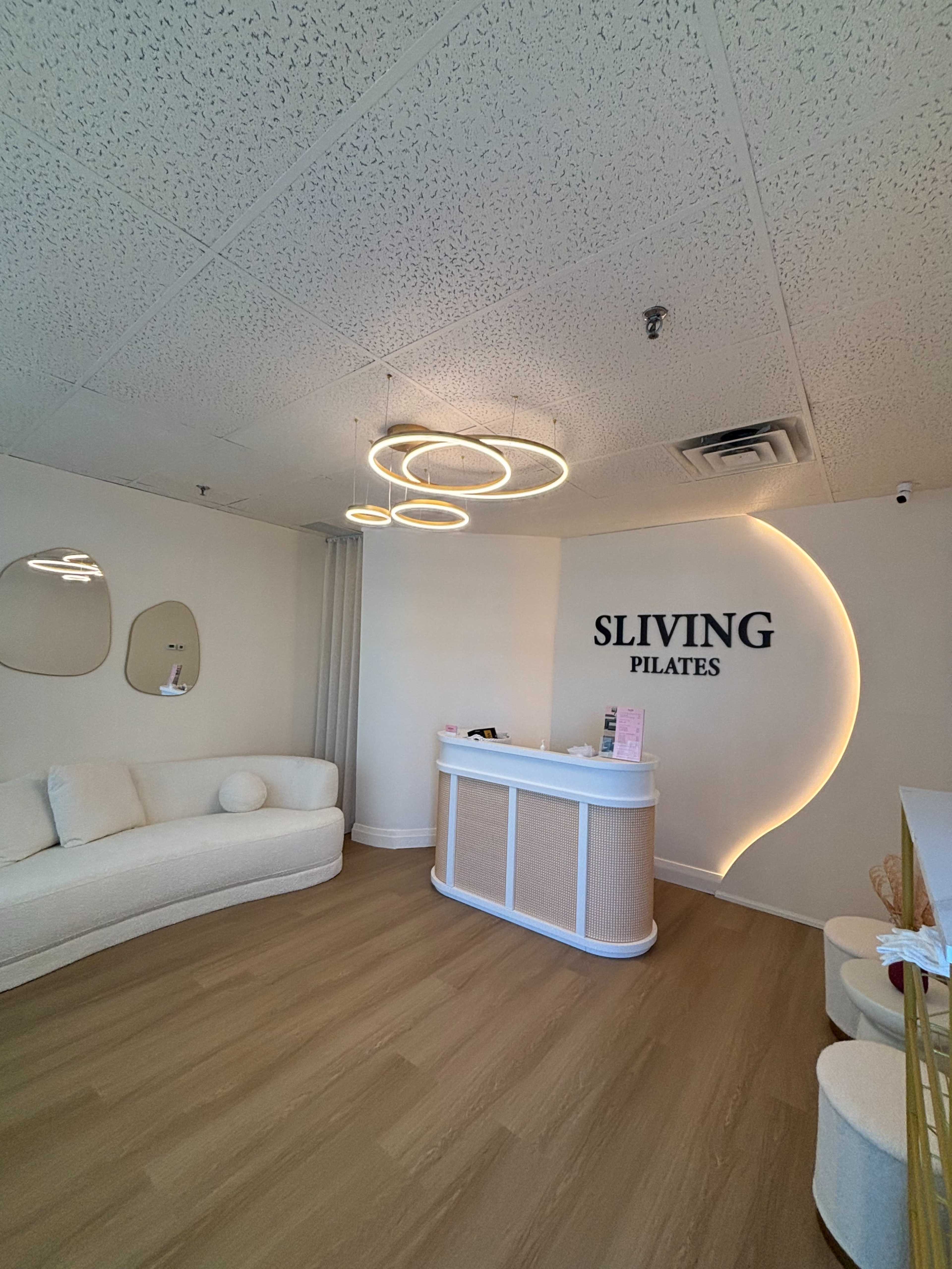 The image shows a modern Pilates studio reception area with a curved front desk, minimalist decor, and soft lighting.