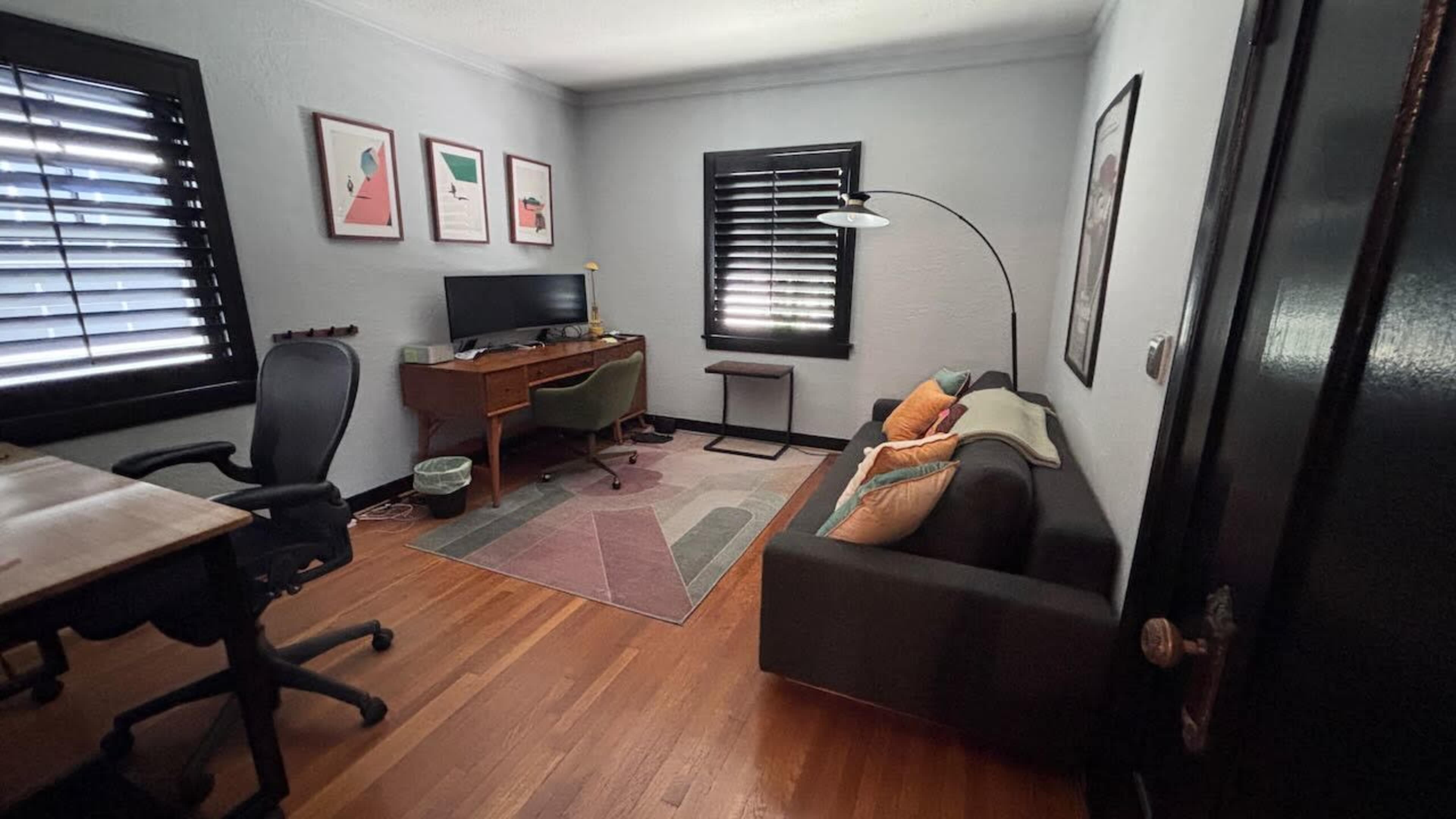 The image shows a small, minimalist office space with a desk, a computer setup, a green chair, and a sofa adorned with cushions.