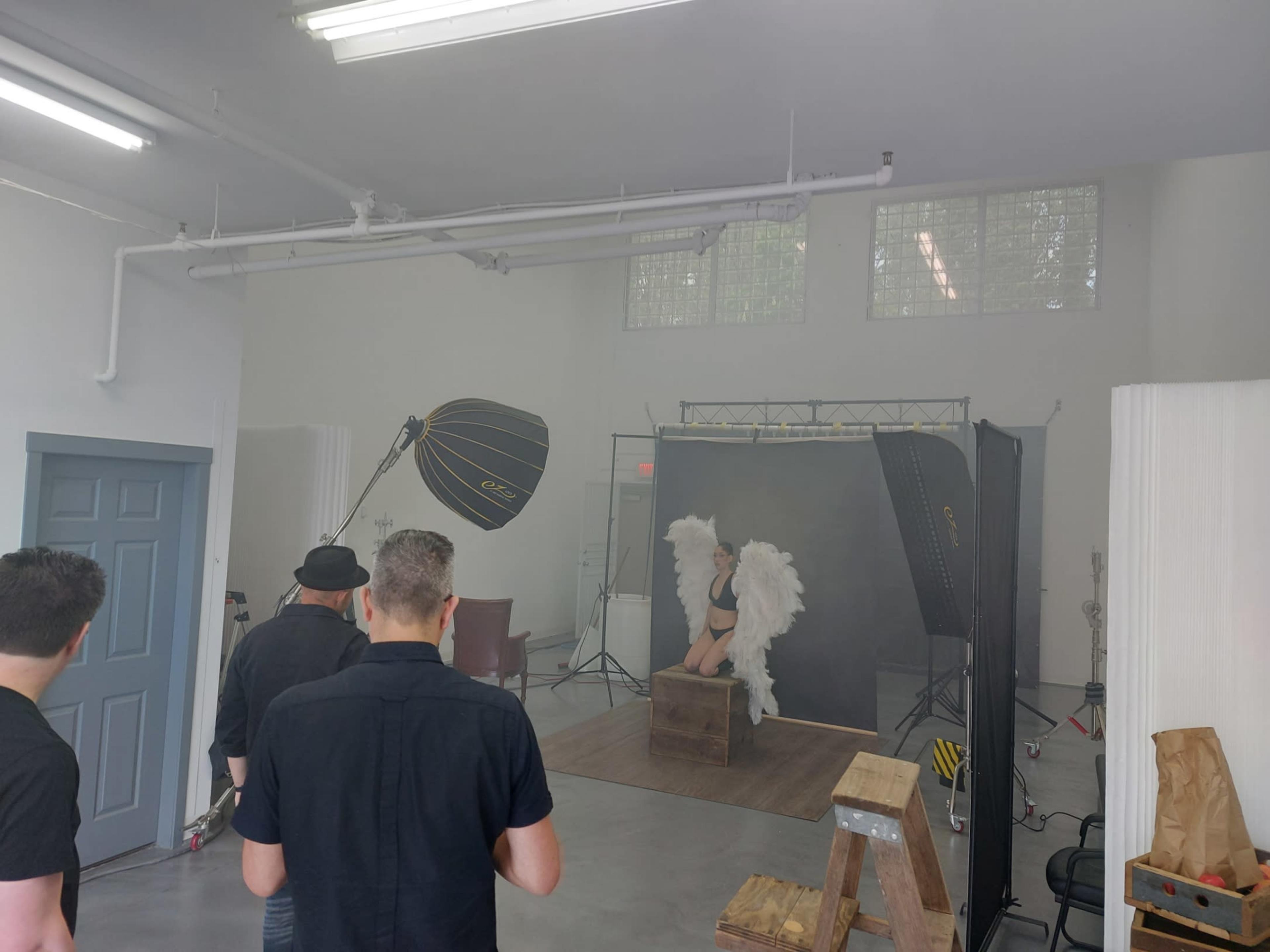 A photoshoot setup features a model with wings posing on a wooden box, while photographers adjust equipment in a spacious studio.