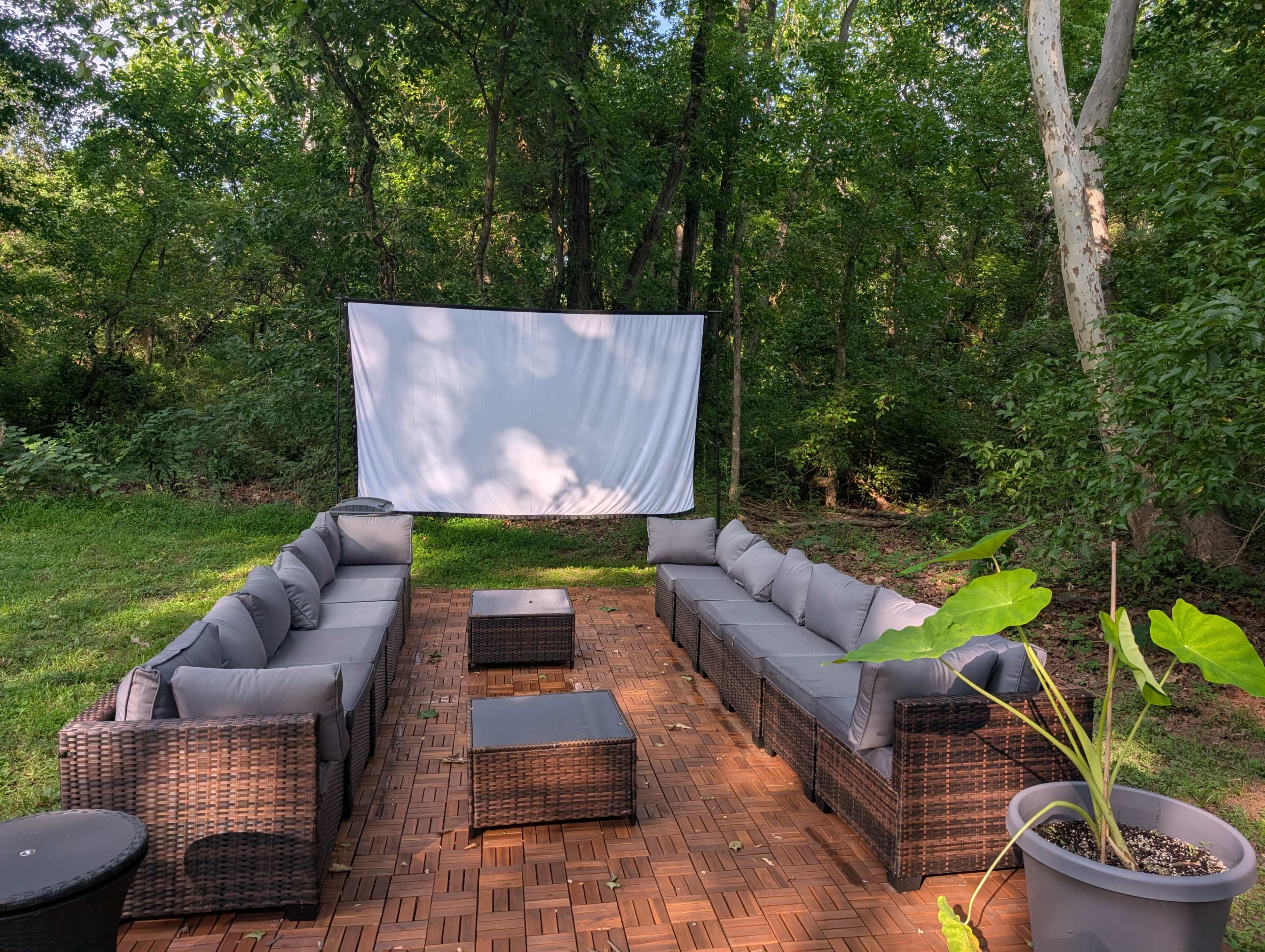 A cozy outdoor seating area with a large white screen set up amidst a green, wooded backdrop.