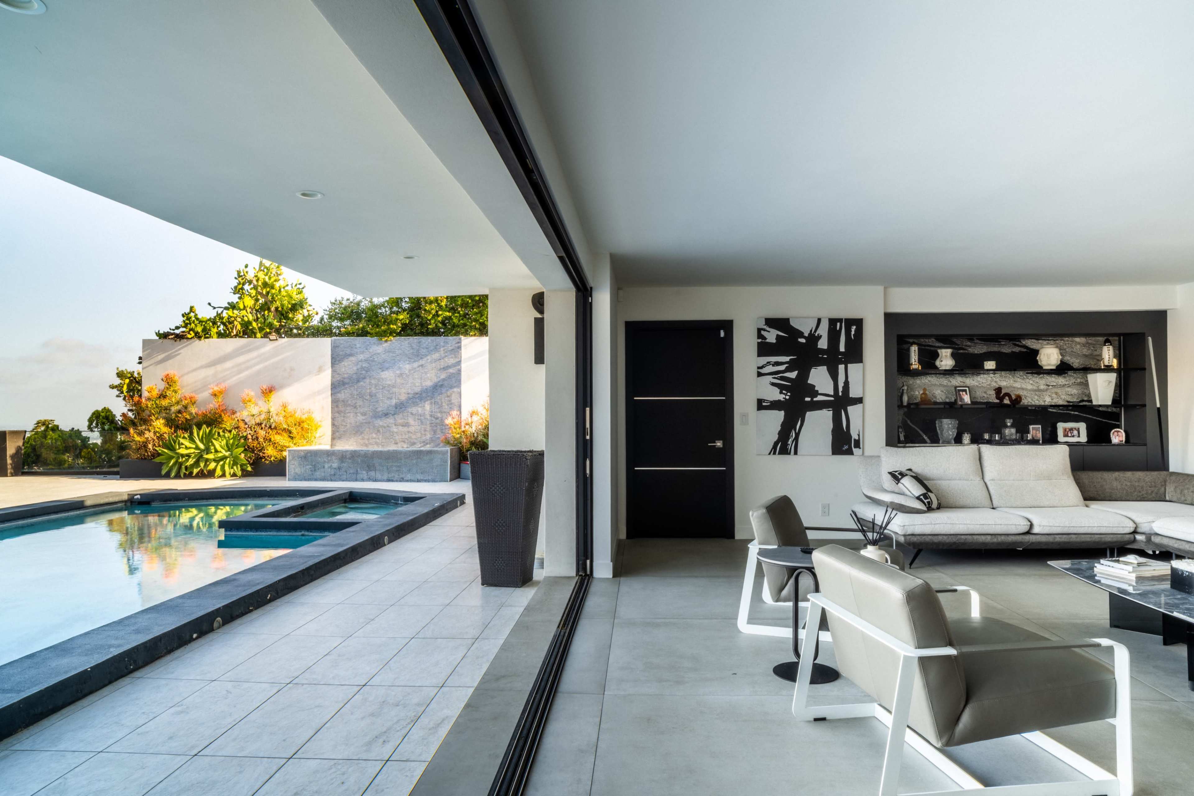 The image shows a modern interior space seamlessly connected to an outdoor pool area through large sliding glass doors.
