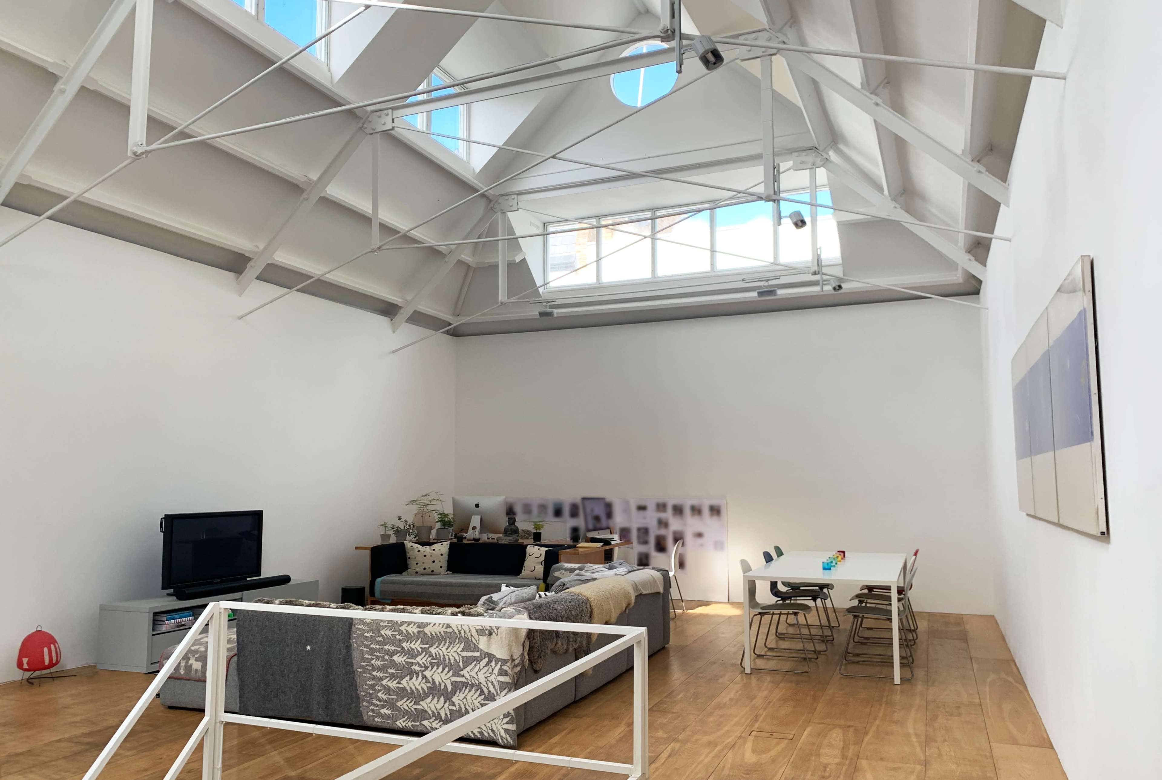 The image shows a spacious, modern living room with a high, vaulted ceiling, a wooden floor, a sectional couch, a television, and a dining table with chairs.