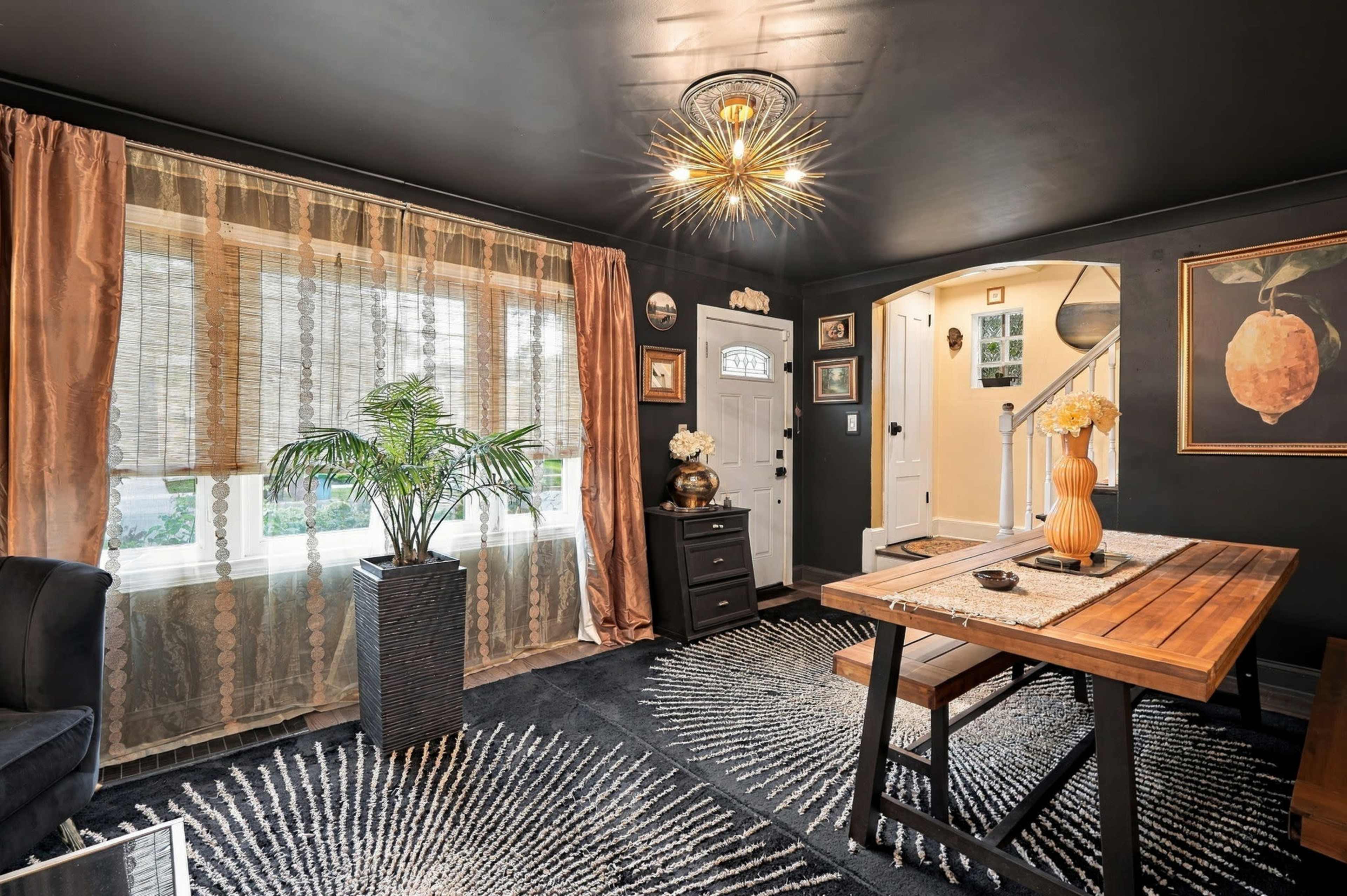 A dining area features a wooden table, black chairs, large windows with patterned curtains, and decorative elements on the walls.
