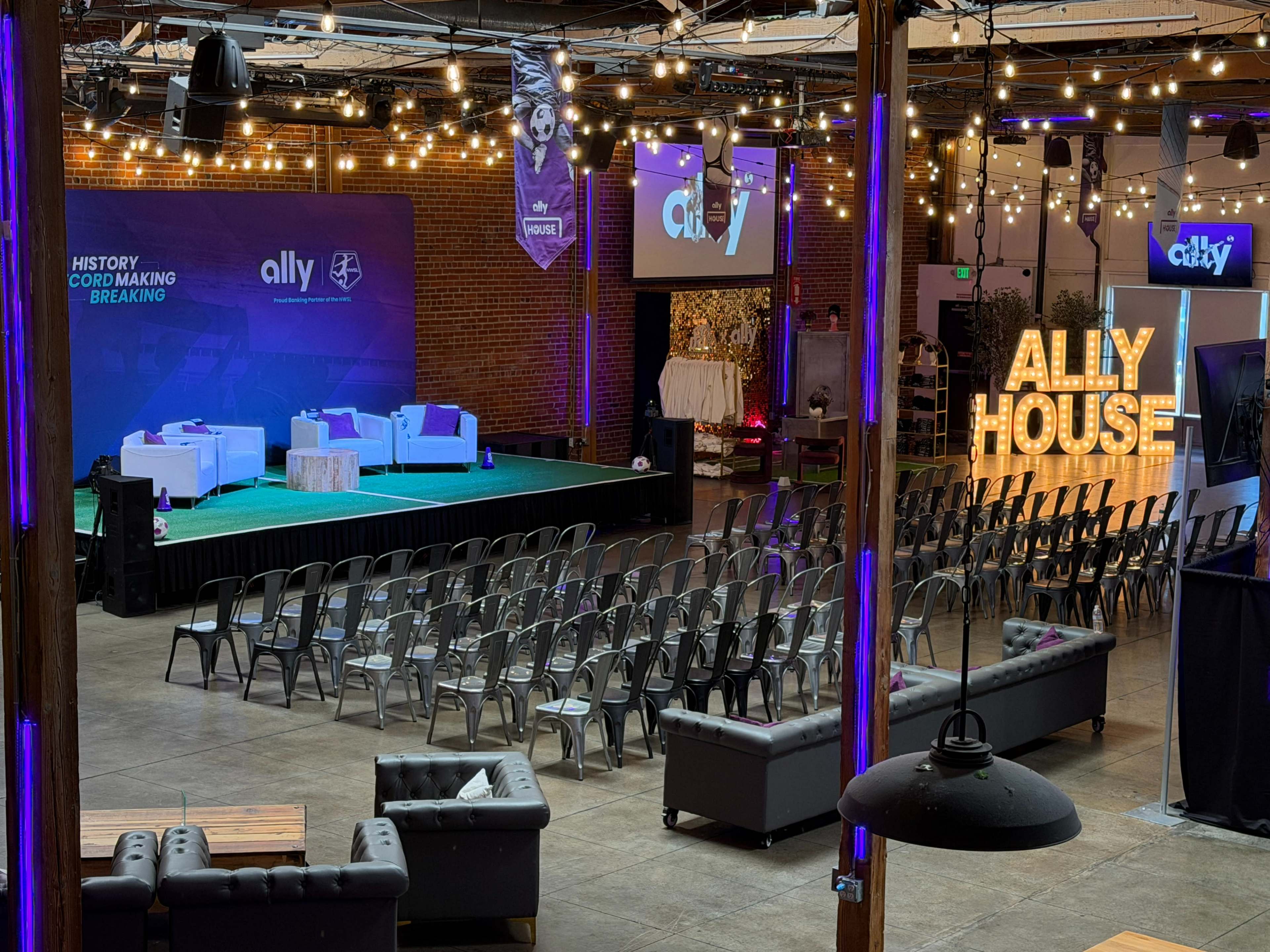 The image shows a spacious event venue with rows of empty chairs facing a stage lit by string lights, featuring banners and a large illuminated sign that reads "ALLY HOUSE."