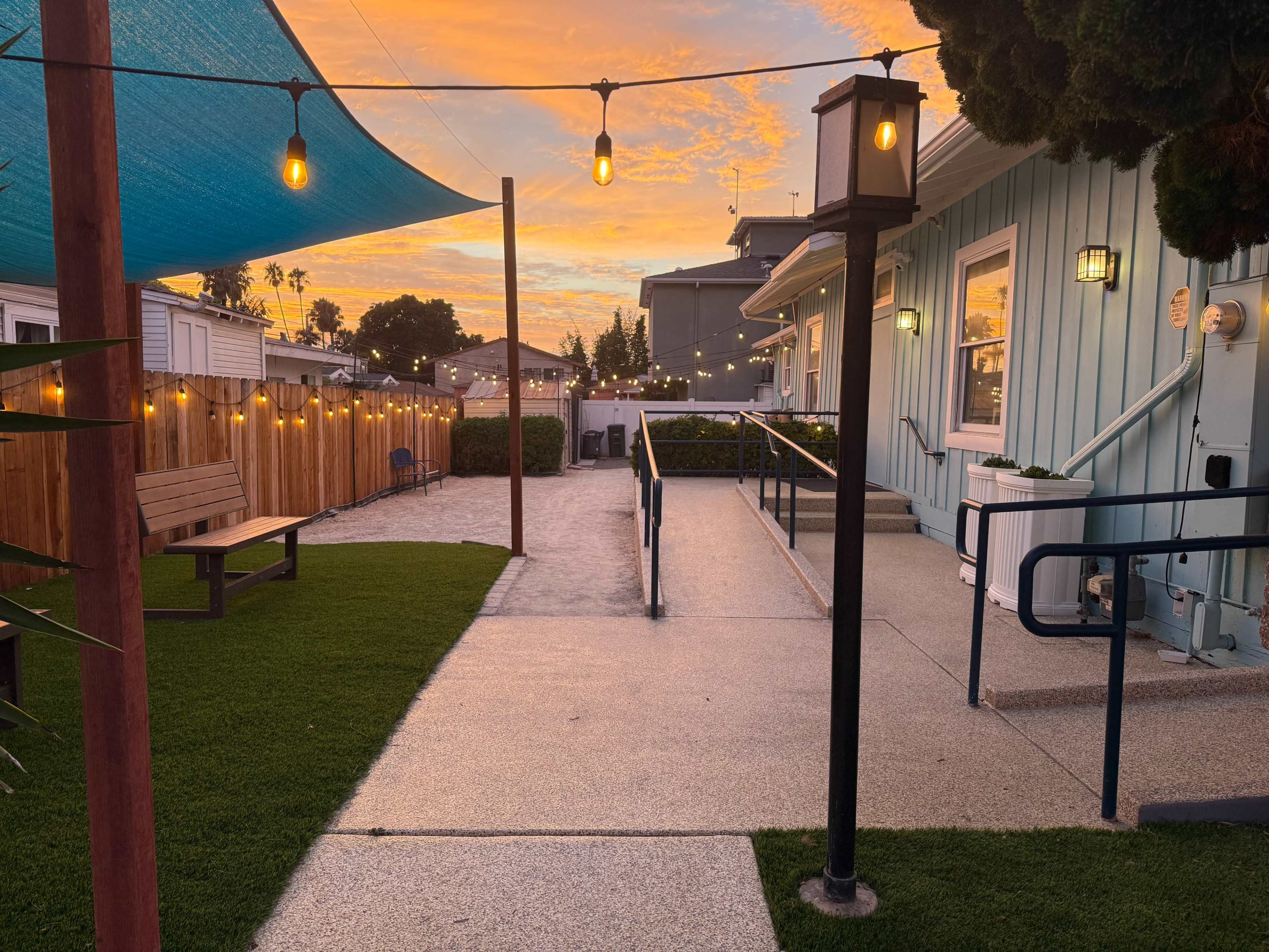 The image shows a patio area with a shaded walkway, benches, and string lights against a vibrant sunset sky.