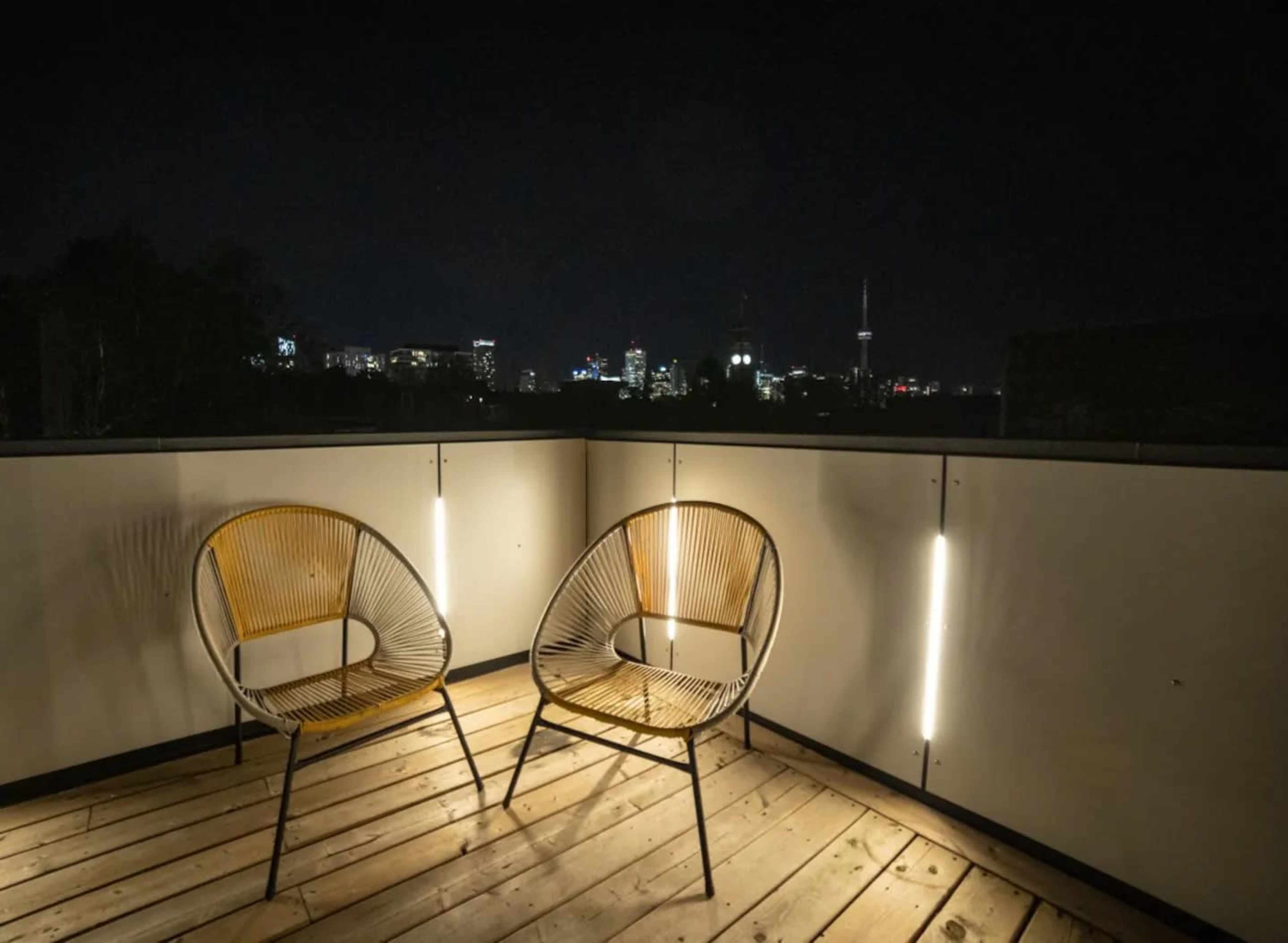 Two woven chairs are positioned on a wooden deck at night, with a city skyline illuminated in the background.