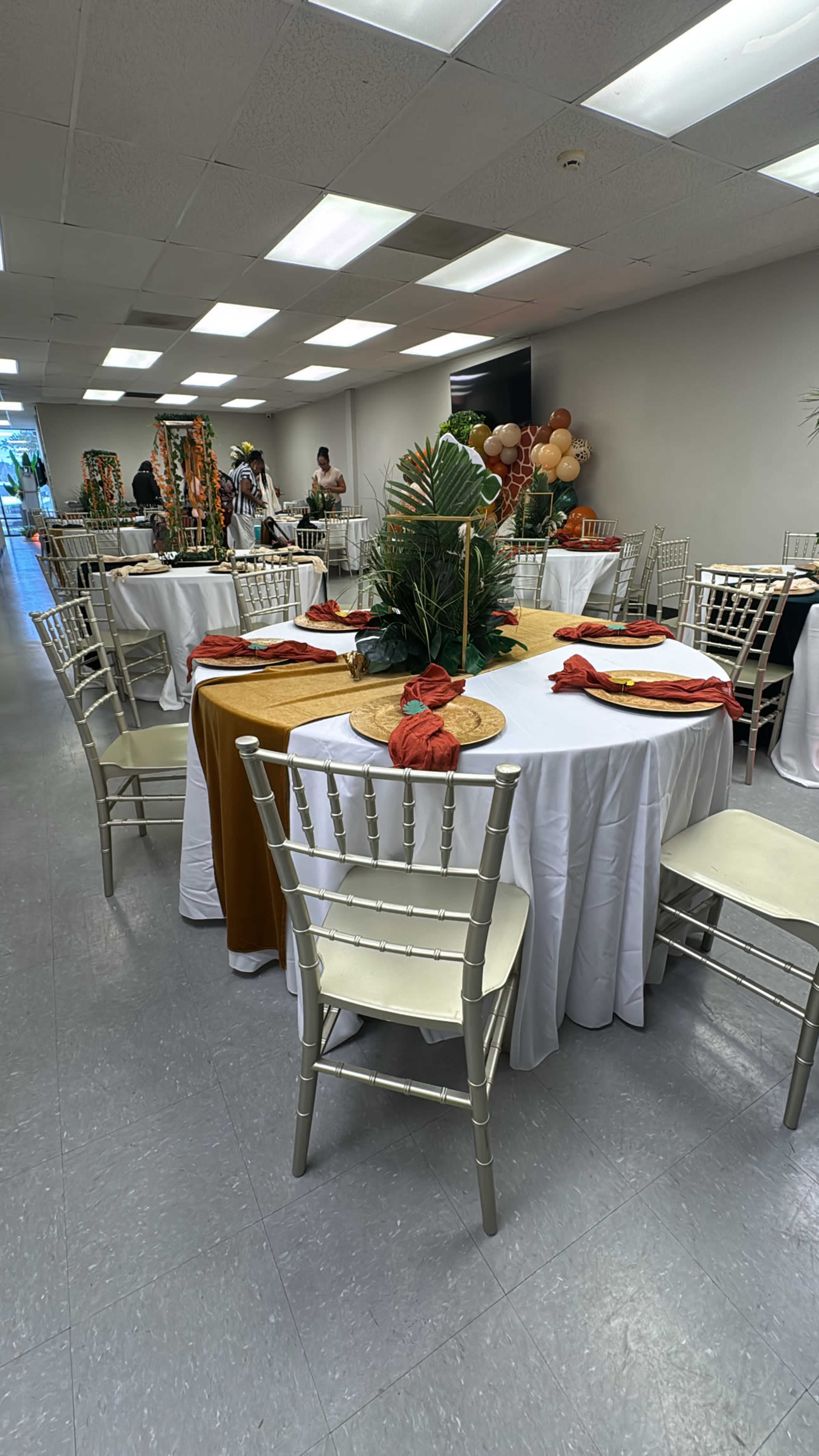 The image shows a banquet hall set for an event, featuring round tables with tablecloths, centerpieces, and chairs arranged neatly.