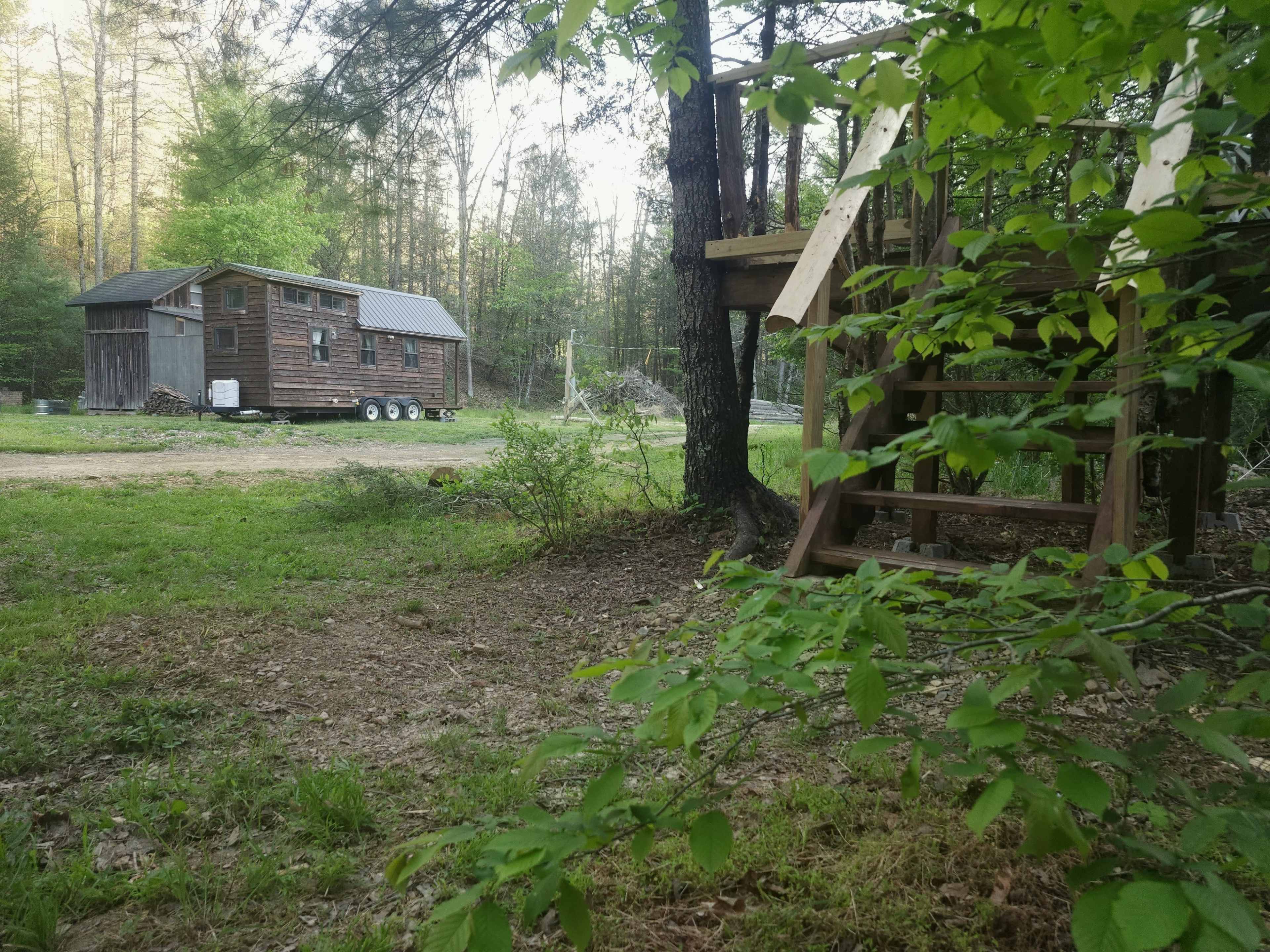 A small wooden cabin on wheels is situated in a wooded area next to a wooden play structure.
