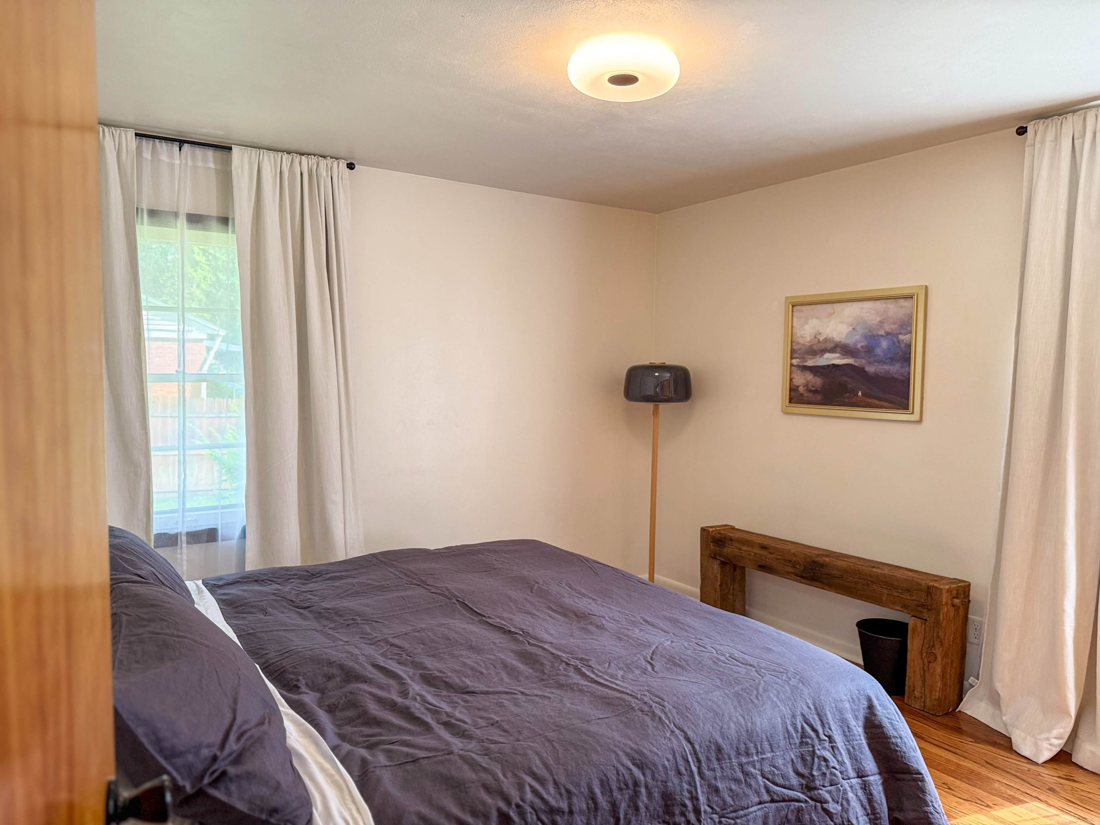 The room features a double bed with dark bedding, a simple wooden bench against the wall, and a lamp beside a window draped with white curtains.