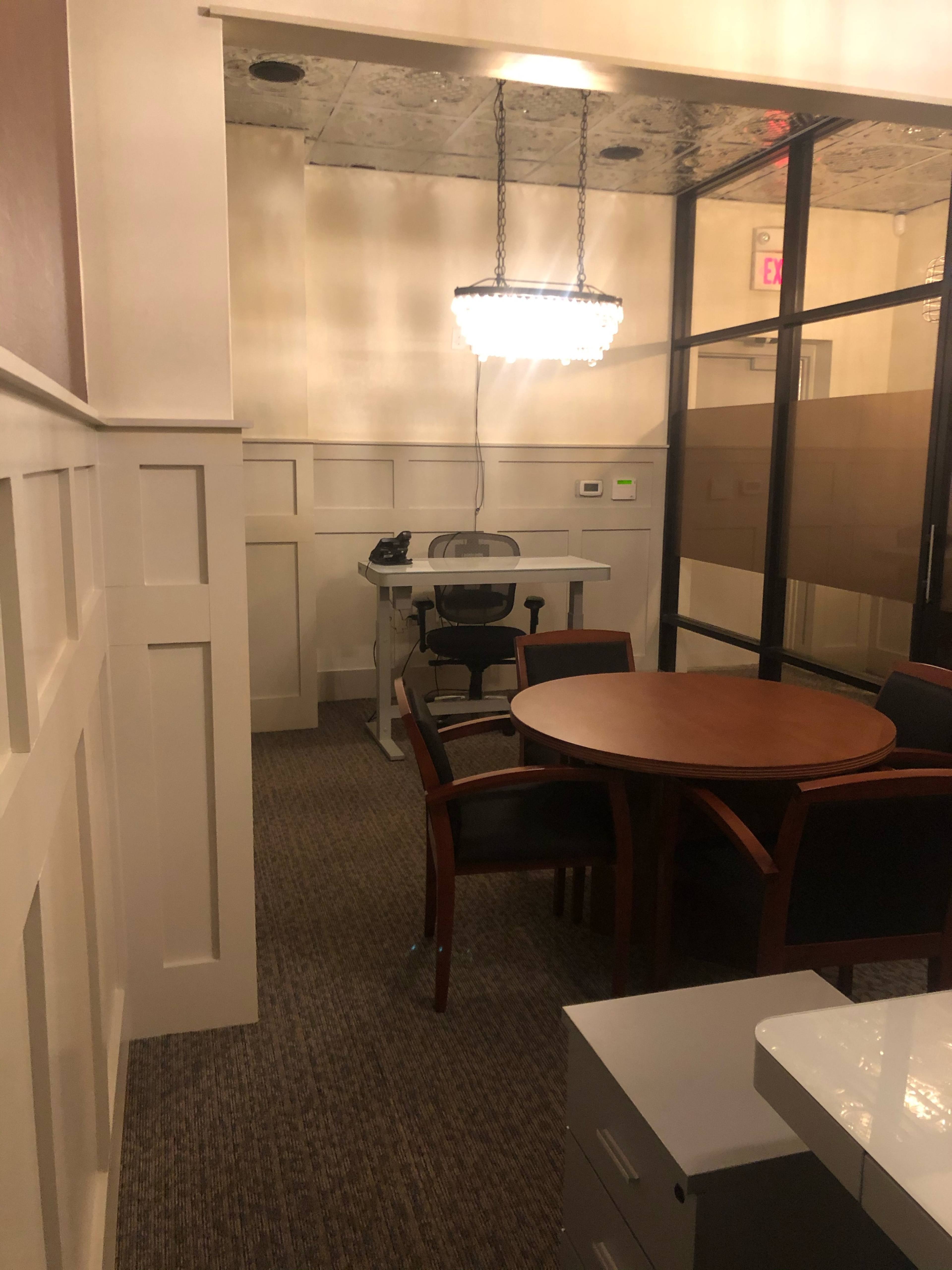 A conference room with a round table and chairs, a desk with an office chair, and a chandelier above.