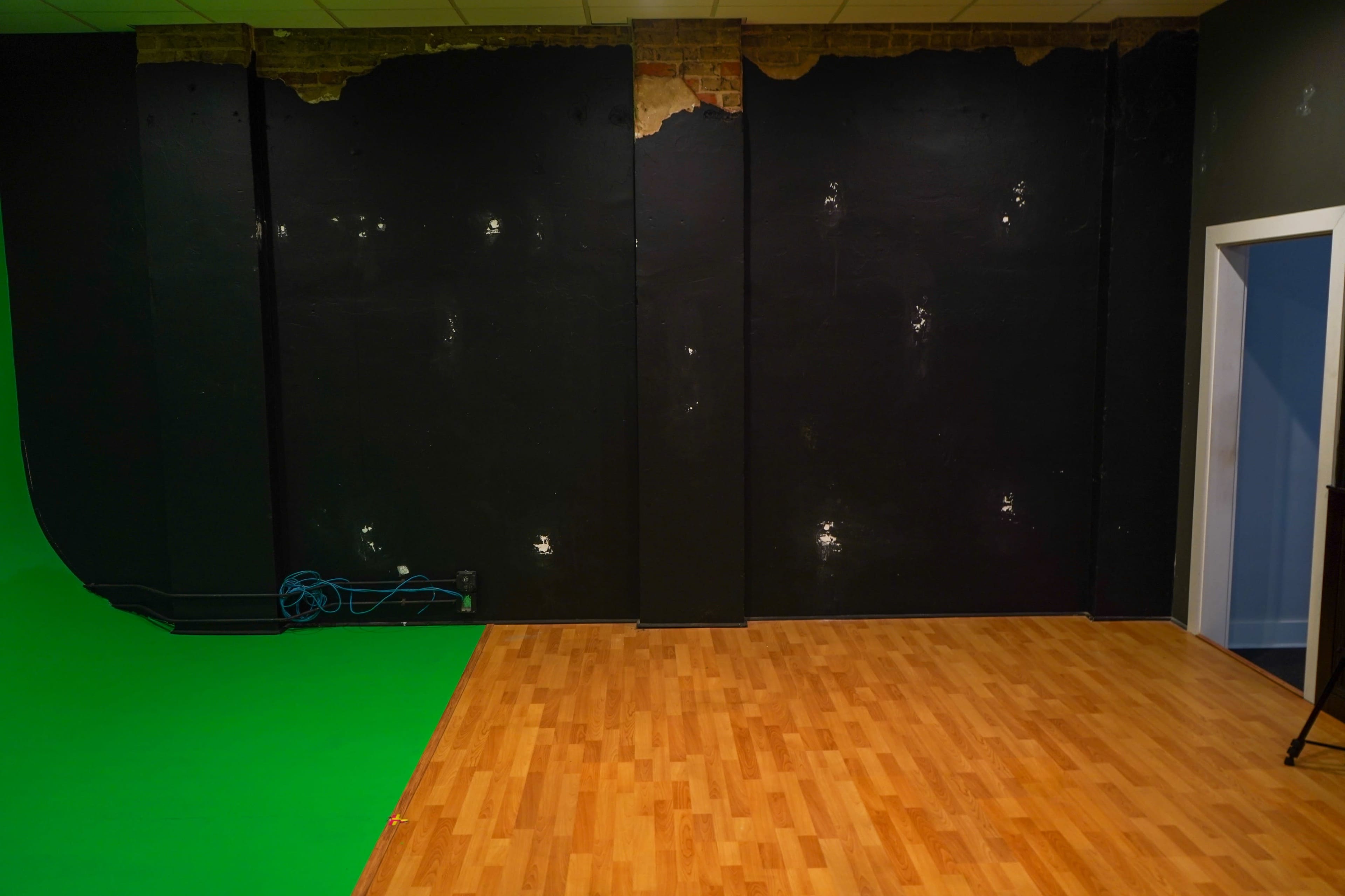 A room with a green screen on one side, black walls with visible damage, and a wooden floor.
