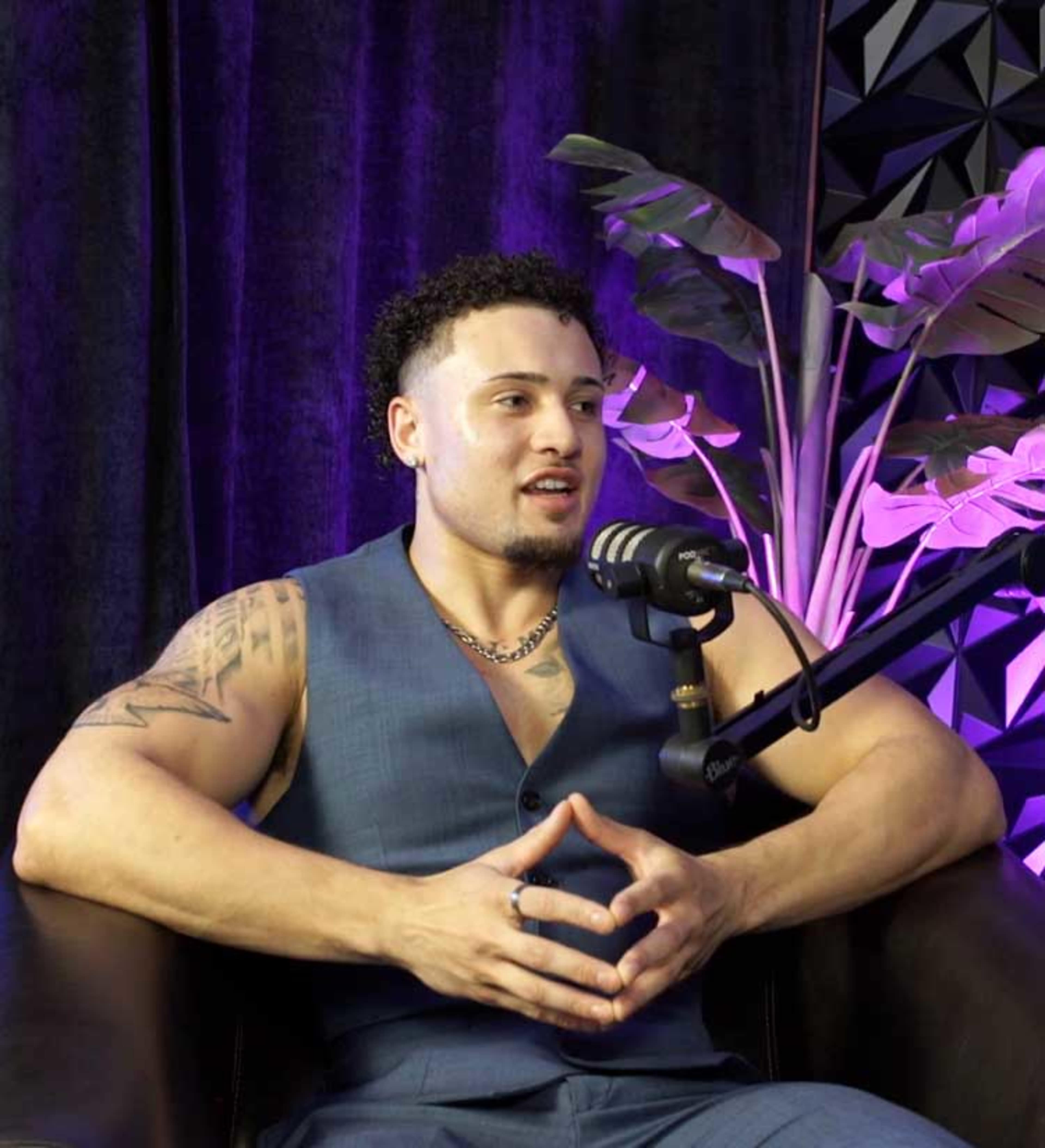 A man with curly hair sits in a chair, wearing a blue vest and speaking into a microphone with plants in the background.